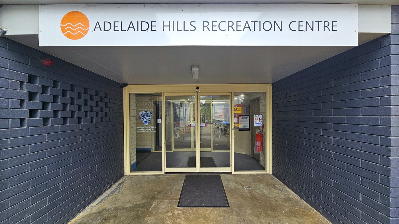 The entrance is enclosed by dark blue brick walls. The door consists of two sets of automatic, glass sliding doors with stickers and posters on the wall, including a fire extinguisher. A non-slippery mat is placed on the doorstep. A distinctive sign, "Adelaide Hills Recreation Centre," is positioned on top near the ceiling.