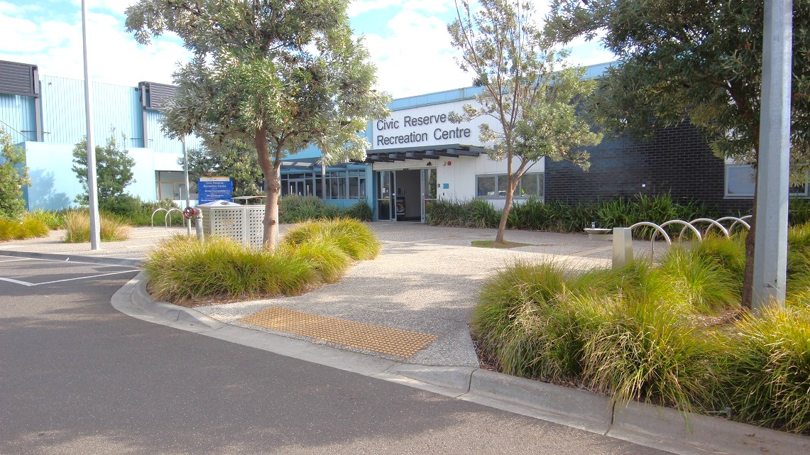 The foreground features a paved area with light-coloured stones and scattered grass patches. On the left, there’s a grey parking space marked with a single parking sign. The entrance of the Civic Reserve Recreation Centre is centrally positioned, featuring large windows and a prominent sign above the door. To the right, there's a landscaped area with shrubs and small trees, along with a decorative garden edging. The foreground features a paved area with light-coloured stones and scattered grass patches. On the left, there’s a grey parking space marked with a single parking sign. The entrance of the Civic Reserve Recreation Centre is centrally positioned, featuring large windows and a prominent sign above the door. To the right, there's a landscaped area with shrubs and small trees, along with a decorative garden edging.