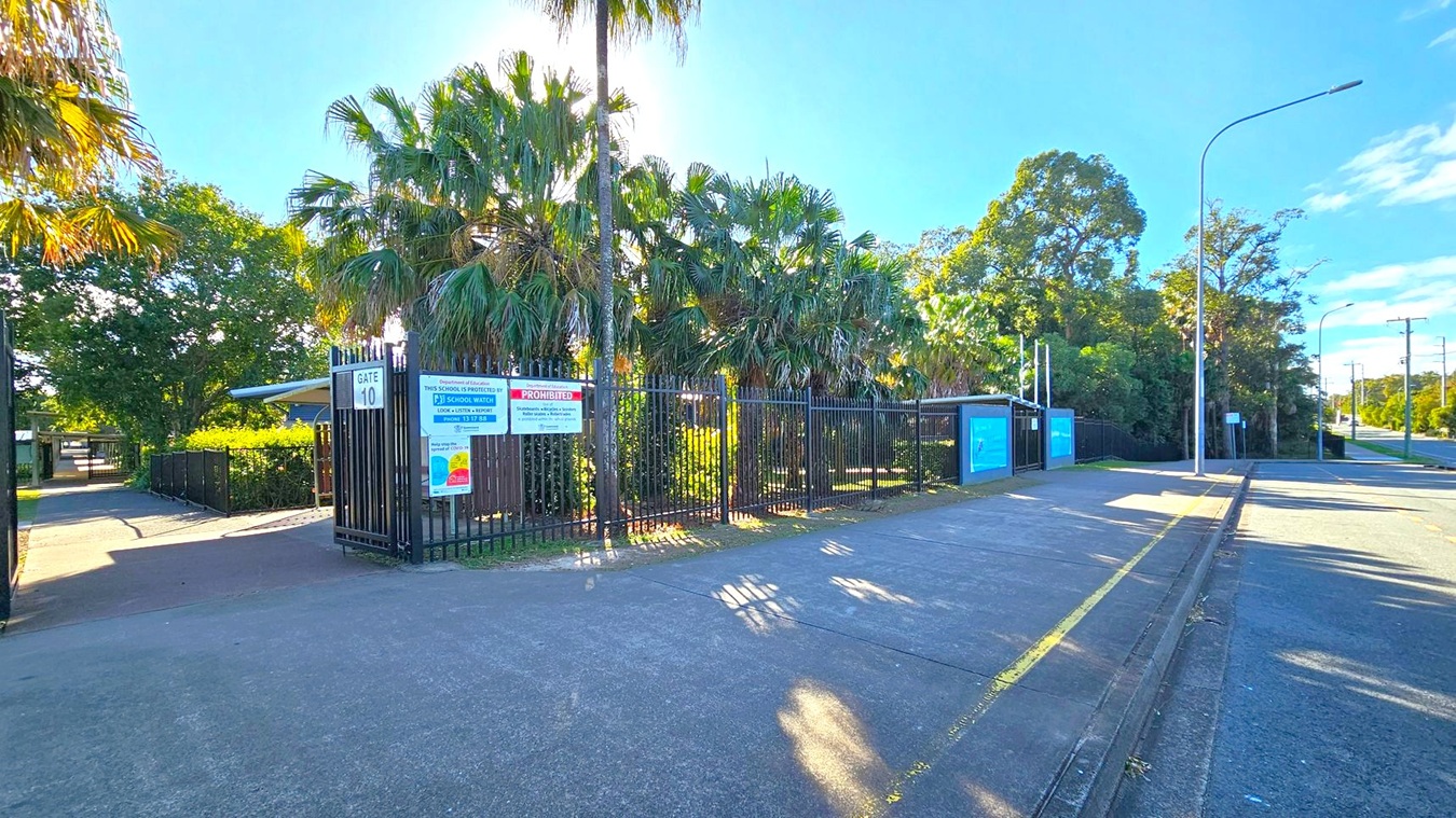 A gated entrance marked "Gate 10" with signs about school security and prohibited activities. Tall palm trees and dense greenery are behind the black metal fence. The pavement is wide, with a yellow line along the kerb. A streetlight and power lines run along the road on the right. The sky is clear and blue, casting shadows on the ground.