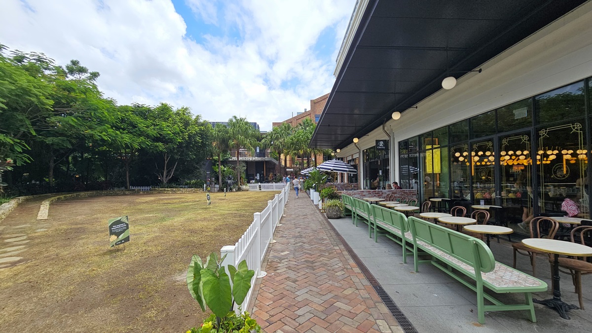 To the left, a field with sparse grass and soil stretches out. Tall trees and decorative shrubs fill a concrete planter. Signs stand near a white fence. On the right, a three-story building with a brick front and large windows is visible. Large striped umbrellas line the front of the restaurant's Al Fresco area, with another brick building adjacent to a taller, multi-story structure behind it. To the left, a field with sparse grass and soil stretches out. Tall trees and decorative shrubs fill a concrete planter. Signs stand near a white fence. On the right, a three-story building with a brick front and large windows is visible. Large striped umbrellas line the front of the restaurant's Al Fresco area, with another brick building adjacent to a taller, multi-story structure behind it.