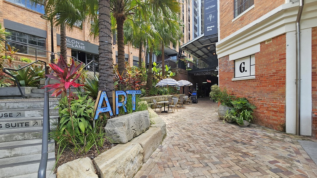 A stairway with handrails leads to a brick-faced building. Plants and palm trees adorn the front, with an "ART" sign. To the right stands another brick building marked "G" with directional signs. On its left, large striped umbrellas shade arranged tables and chairs. Further back, a shaded area with ornamental plants against the wall. The brick floor extends to the rear. A stairway with handrails leads to a brick-faced building. Plants and palm trees adorn the front, with an "ART" sign. To the right stands another brick building marked "G" with directional signs. On its left, large striped umbrellas shade arranged tables and chairs. Further back, a shaded area with ornamental plants against the wall. The brick floor extends to the rear.