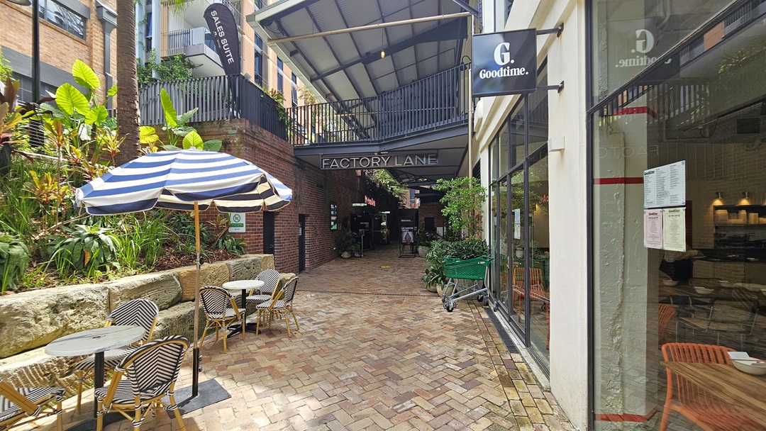 On the left, ornamental plants and palm trees line the landscape, bordered by large rocks. Nearby, a massive striped umbrella shade arranged tables and chairs. To the right, a glass wall reveals the restaurant inside the building. The brick pathway leads towards the shaded area of a red brick building. A green trolley sits near the window, accompanied by ornamental plants in large pots. On the left, ornamental plants and palm trees line the landscape, bordered by large rocks. Nearby, a massive striped umbrella shade arranged tables and chairs. To the right, a glass wall reveals the restaurant inside the building. The brick pathway leads towards the shaded area of a red brick building. A green trolley sits near the window, accompanied by ornamental plants in large pots.