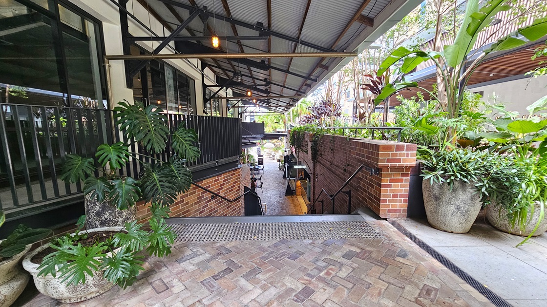 A staircase with handrails descends, preceded by truncated domes. Large pots of ornamental plants adorn the front and right sides. The left building features a corrugated roof extending to the right, secured by a low black metal fence. Brick walls flank both sides. Below, tables and chairs are visible. A staircase with handrails descends, preceded by truncated domes. Large pots of ornamental plants adorn the front and right sides. The left building features a corrugated roof extending to the right, secured by a low black metal fence. Brick walls flank both sides. Below, tables and chairs are visible.