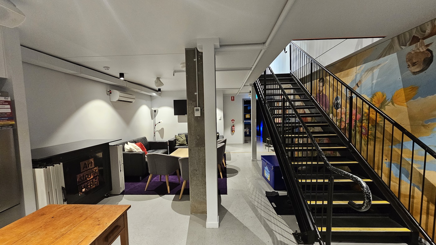 To the right, there's a staircase with handrails, under which is a blue cooler box. To the left, there's a seating area with cushioned chairs and a wooden table. More wooden tables are positioned near the wall and fridge. A large colourful painting fills the entire wall on the right. The floor, walls, and ceilings are white vinyl, equipped with LED lights. To the right, there's a staircase with handrails, under which is a blue cooler box. To the left, there's a seating area with cushioned chairs and a wooden table. More wooden tables are positioned near the wall and fridge. A large colourful painting fills the entire wall on the right. The floor, walls, and ceilings are white vinyl, equipped with LED lights.