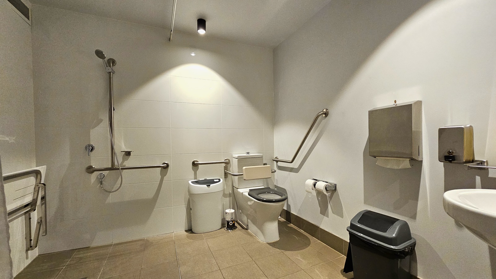 To the left, a foldable bench chair is affixed to the wall. Next to it is a shower with handrails, followed by a toilet bowl with hand grabs. A rubbish bin and tissue dispenser are beside it. To the right, there's a washbasin, another tissue dispenser, and a soap dispenser mounted on the wall. Underneath is another rubbish bin. The floor has cream tiles, while the walls and ceiling are white. To the left, a foldable bench chair is affixed to the wall. Next to it is a shower with handrails, followed by a toilet bowl with hand grabs. A rubbish bin and tissue dispenser are beside it. To the right, there's a washbasin, another tissue dispenser, and a soap dispenser mounted on the wall. Underneath is another rubbish bin. The floor has cream tiles, while the walls and ceiling are white.