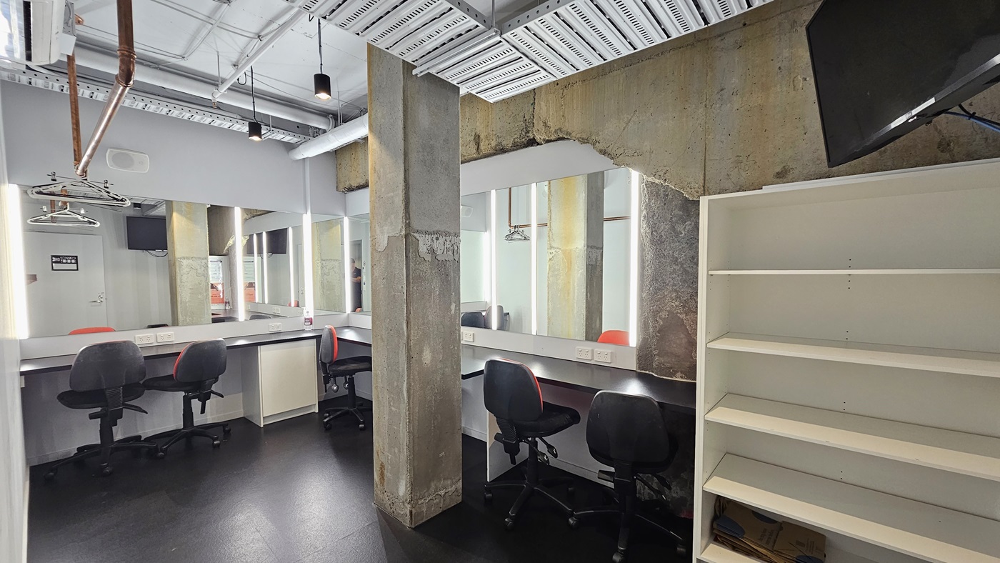 The dressing room features mirrors surrounding the walls. Adjustable chairs are positioned near the mirrors. To the right, there's a white display rack, with a flatscreen mounted on top. The ceiling displays exposed concrete beams, pipes, and air conditioning vents. The walls are white, and the floor is tiled black. The dressing room features mirrors surrounding the walls. Adjustable chairs are positioned near the mirrors. To the right, there's a white display rack, with a flatscreen mounted on top. The ceiling displays exposed concrete beams, pipes, and air conditioning vents. The walls are white, and the floor is tiled black.