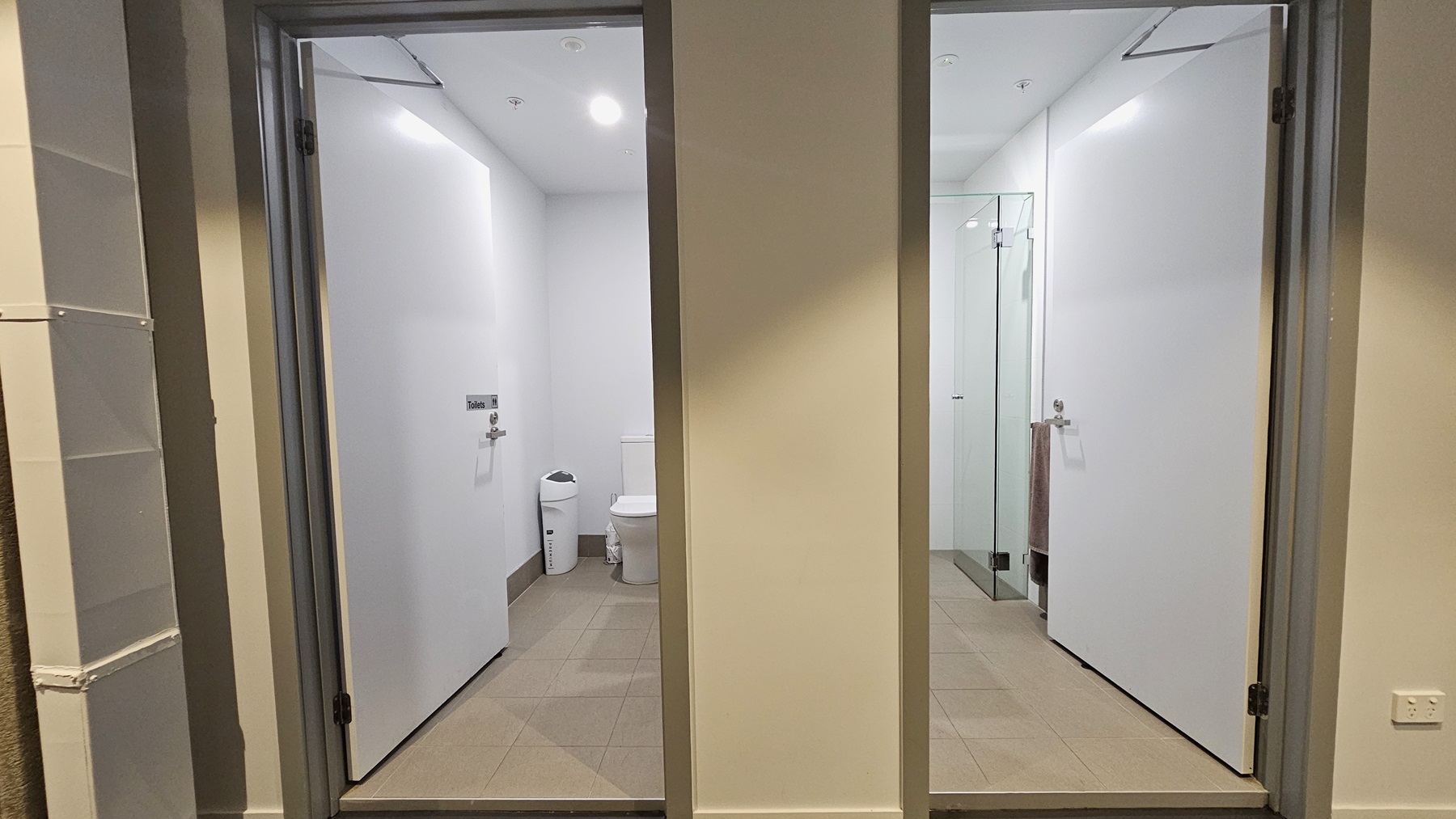 Two inward-opening doors. The first reveals a toilet bowl placed near the corner, accompanied by a white rubbish bin. The second door unveils a glass-enclosed shower area. The floor is tiled with brown, and the walls and ceiling are all white with LED lights. Two inward-opening doors. The first reveals a toilet bowl placed near the corner, accompanied by a white rubbish bin. The second door unveils a glass-enclosed shower area. The floor is tiled with brown, and the walls and ceiling are all white with LED lights.