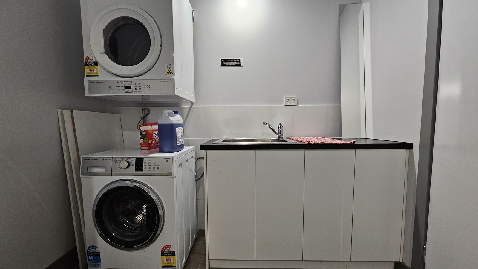 A laundry machine with detergent soaps on top. Another laundry machine is overhead. To the right, there's a small sink with a faucet, and a kitchen towel placed on top. The walls are all white, and the floor has brown tiles. An instruction sign is placed on the wall. A laundry machine with detergent soaps on top. Another laundry machine is overhead. To the right, there's a small sink with a faucet, and a kitchen towel placed on top. The walls are all white, and the floor has brown tiles. An instruction sign is placed on the wall.