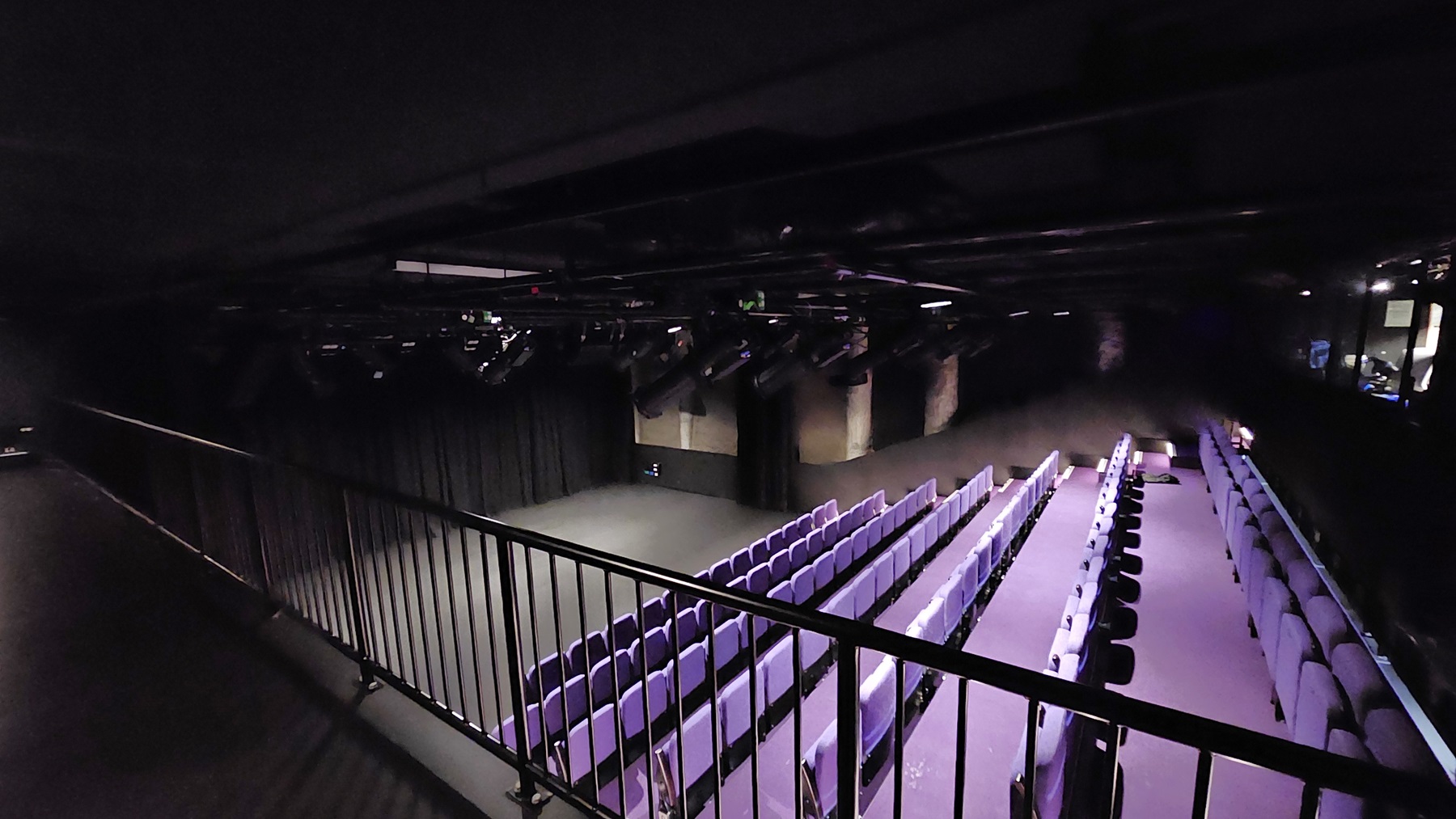 A dimly lit area with black floor tiles and walls. Metal railing secures the space. Overlooking below is the stage with black tiles, surrounded by black walls and curtains. Adjacent to the stage is tiered seating with lavender chairs. The ceiling features exposed pipes, audio equipment, and lighting fixtures. A dimly lit area with black floor tiles and walls. Metal railing secures the space. Overlooking below is the stage with black tiles, surrounded by black walls and curtains. Adjacent to the stage is tiered seating with lavender chairs. The ceiling features exposed pipes, audio equipment, and lighting fixtures.