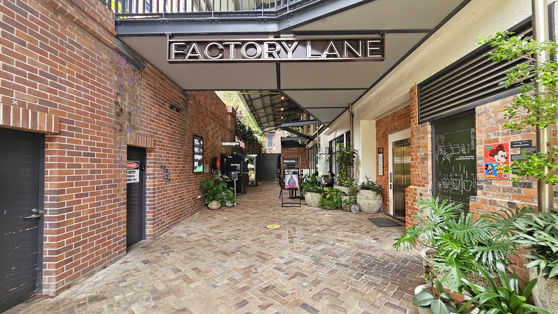 Both sides of the building display a red brick facade with black doors. A ceiling-hung signage reads "Factory Lane". On the right, a lift is positioned. Large pots showcase ornamental plants upfront, alongside free standing banners. Posters adorn the brick walls, with a concrete staircase visible further ahead. Both sides of the building display a red brick facade with black doors. A ceiling-hung signage reads "Factory Lane". On the right, a lift is positioned. Large pots showcase ornamental plants upfront, alongside free standing banners. Posters adorn the brick walls, with a concrete staircase visible further ahead.