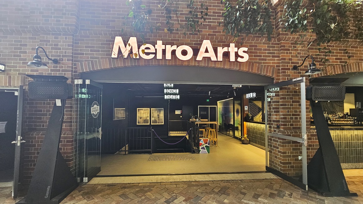 On the left stands a building with a brick facade labelled "Metro Arts". Ornamental plants hang above, extending towards the building. Lighting fixtures adorn the wall, with two black speaker-like structures positioned in front of the glass door entrance. Inside, a staircase with handrails is equipped with truncated domes and a rope bollard. To the right, a bar is arranged with movable chairs for seating. On the left stands a building with a brick facade labelled "Metro Arts". Ornamental plants hang above, extending towards the building. Lighting fixtures adorn the wall, with two black speaker-like structures positioned in front of the glass door entrance. Inside, a staircase with handrails is equipped with truncated domes and a rope bollard. To the right, a bar is arranged with movable chairs for seating.