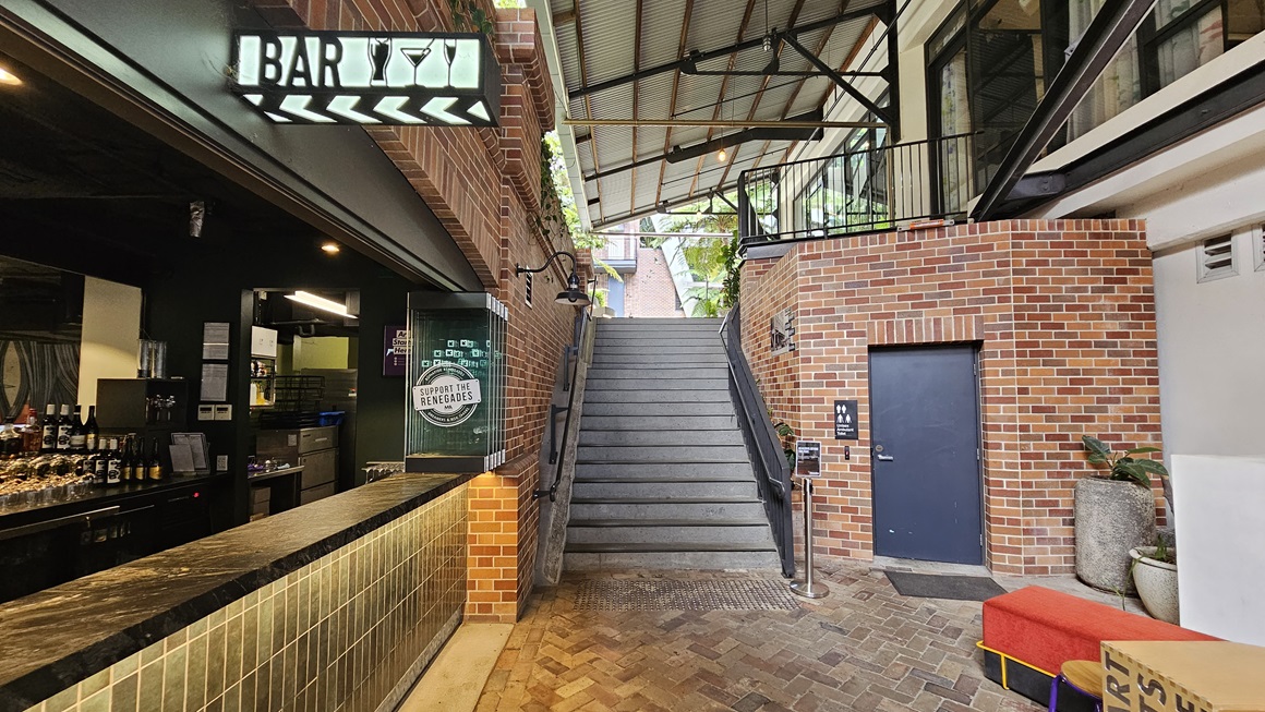To the left stands a building with a brick facade, with a neon sign reading "Bar". Inside, a brick wall decorates the bar area, with wines and liquors displayed on the counter. A staircase equipped with handrails and truncated domes is positioned in the middle. To the right, an accessible toilet with a brick facade and grey door is situated. Ornamental plants in large pots adorn the right side, while the corrugated roof extends to the left, providing shade. Brick tiles cover the floor. To the left stands a building with a brick facade, with a neon sign reading "Bar". Inside, a brick wall decorates the bar area, with wines and liquors displayed on the counter. A staircase equipped with handrails and truncated domes is positioned in the middle. To the right, an accessible toilet with a brick facade and grey door is situated. Ornamental plants in large pots adorn the right side, while the corrugated roof extends to the left, providing shade. Brick tiles cover the floor.