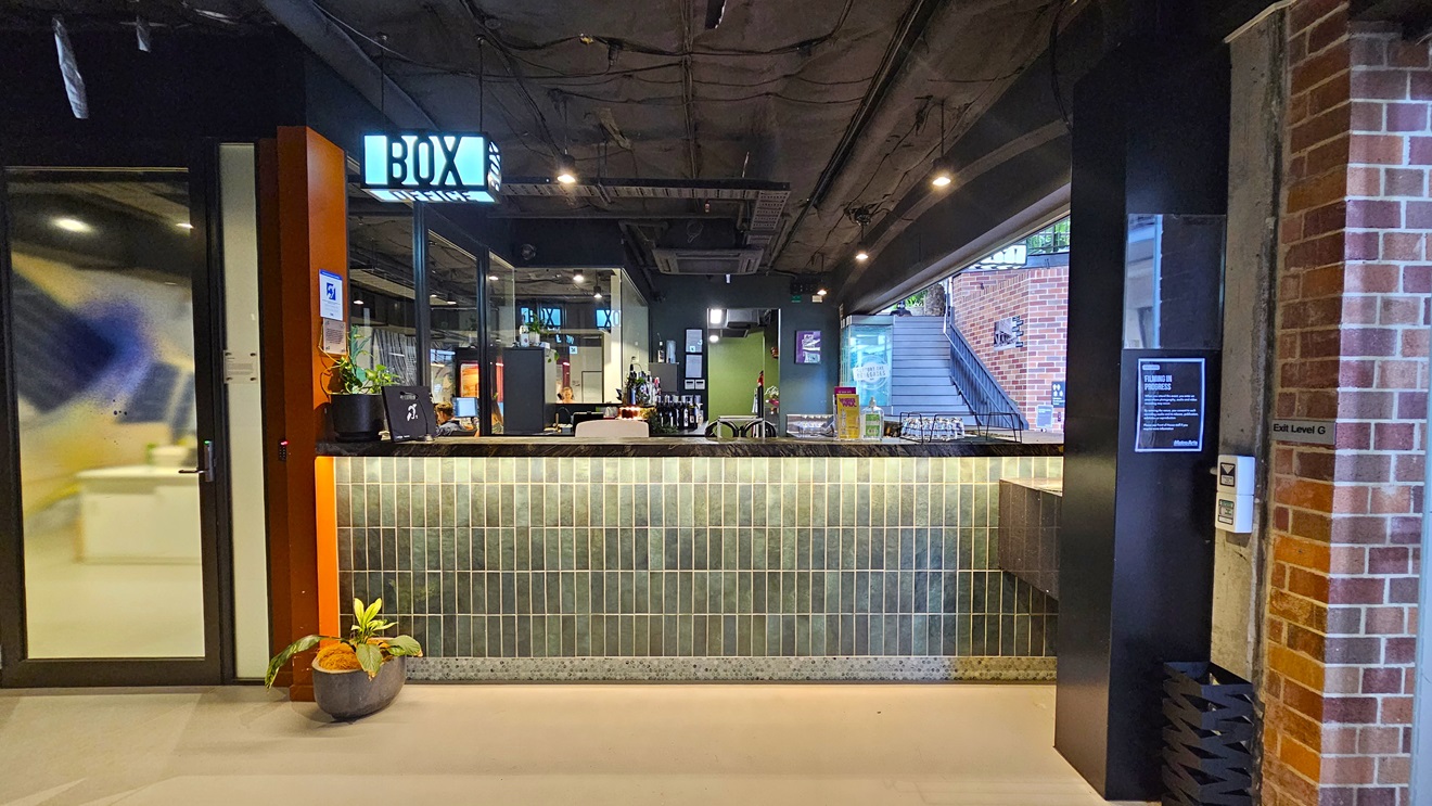 The bar showcases a brick facade with a neon sign reading "Box Office". Wines and liquors are displayed on the counter. A glass door and a pot of ornamental plants adorn the left side. A staircase with handrails and truncated domes is positioned to the right. Signages are affixed to the wall. The ceiling is all black with lights and aircon vents, while the floor is tiled white. The bar showcases a brick facade with a neon sign reading "Box Office". Wines and liquors are displayed on the counter. A glass door and a pot of ornamental plants adorn the left side. A staircase with handrails and truncated domes is positioned to the right. Signages are affixed to the wall. The ceiling is all black with lights and aircon vents, while the floor is tiled white.