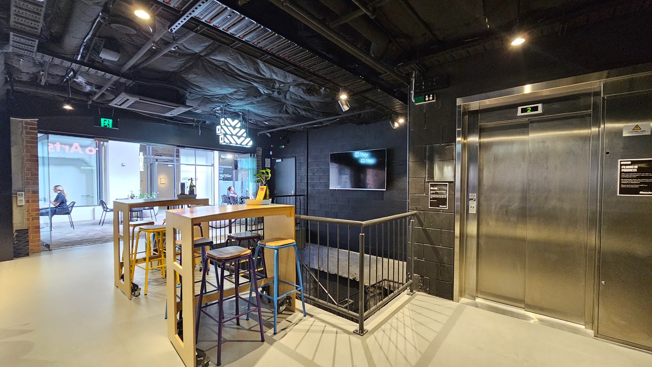 To the right, a lift displays a poster on its wall, adjacent to a flatscreen attached to the black wall. In the middle, two tables with stools are situated, while a staircase leading downwards stands behind. Metal railing secures the area from the outside. The ceiling is all black with lights and aircon vents, and the floor is tiled white. To the right, a lift displays a poster on its wall, adjacent to a flatscreen attached to the black wall. In the middle, two tables with stools are situated, while a staircase leading downwards stands behind. Metal railing secures the area from the outside. The ceiling is all black with lights and aircon vents, and the floor is tiled white.