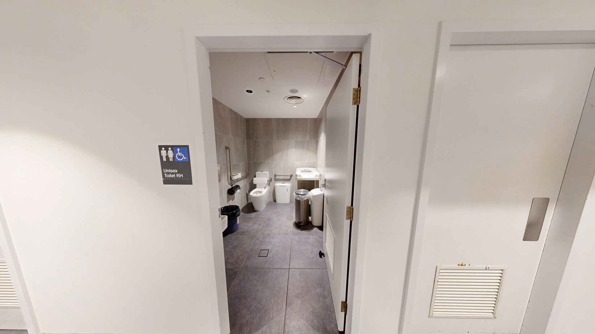 In the unisex bathroom, a toilet bowl is in the left corner with handrails and a tissue dispenser on the wall. A washbasin is positioned to the right alongside rubbish bins. Grey tiles cover the floor and walls. The white ceiling is illuminated by LED lights. Outside, signage indicates all-gender access on the white wall next to another white door. In the unisex bathroom, a toilet bowl is in the left corner with handrails and a tissue dispenser on the wall. A washbasin is positioned to the right alongside rubbish bins. Grey tiles cover the floor and walls. The white ceiling is illuminated by LED lights. Outside, signage indicates all-gender access on the white wall next to another white door.