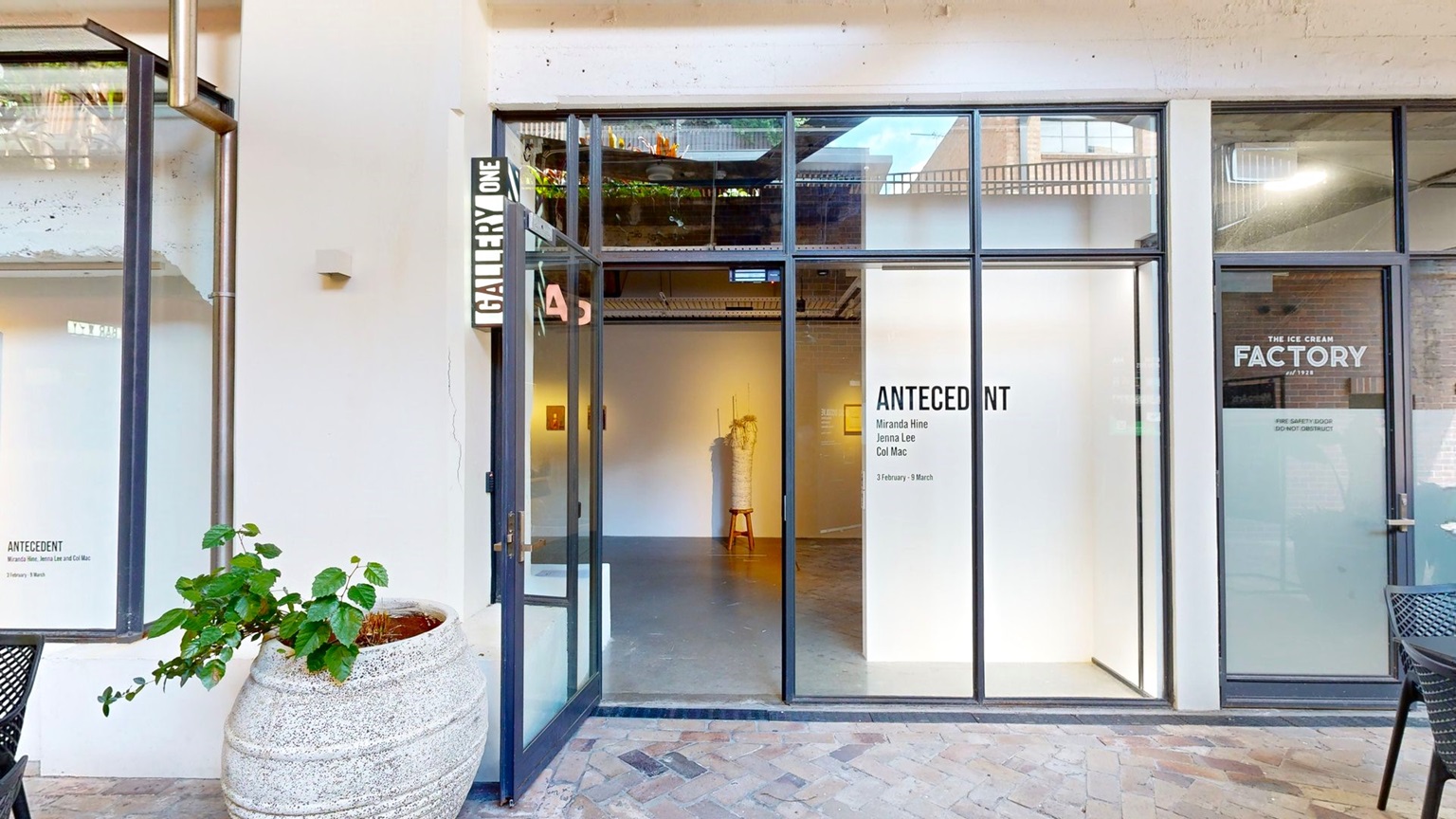 A manual glass door opens outward, with a signage reading "Gallery One" on the wall above. An ornamental plant sits in a huge pot below. Signages are affixed to the right side of the glass door. Movable black tables and chairs are arranged in front. The floor is tiled with brick, and the wall is white. Inside, a section of the gallery is visible. A manual glass door opens outward, with a signage reading "Gallery One" on the wall above. An ornamental plant sits in a huge pot below. Signages are affixed to the right side of the glass door. Movable black tables and chairs are arranged in front. The floor is tiled with brick, and the wall is white. Inside, a section of the gallery is visible.