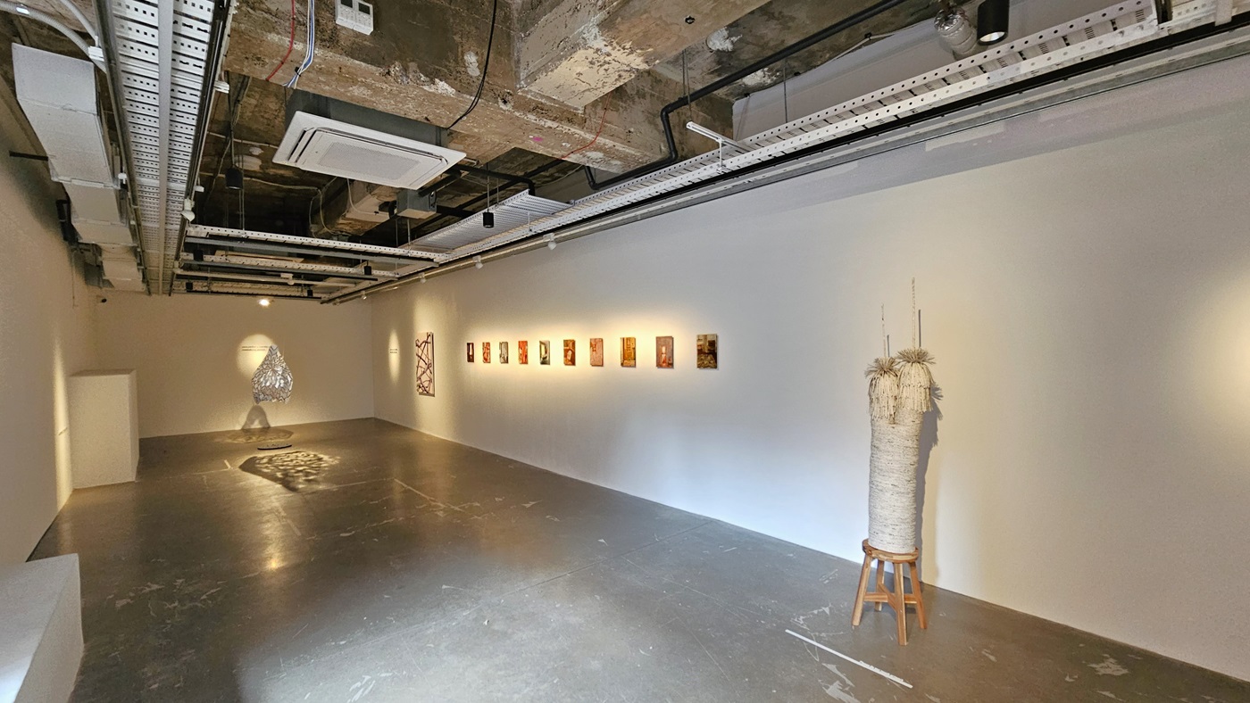 To the right, art paintings adorn the white wall, accompanied by hanging lighting fixtures. A piece of craft made of ropes and strings is displayed against the wall. The ceiling reveals exposed concrete beams, pipes, air conditioning vents, and pin lights. The polished cement floor exhibits worn-out markings. To the right, art paintings adorn the white wall, accompanied by hanging lighting fixtures. A piece of craft made of ropes and strings is displayed against the wall. The ceiling reveals exposed concrete beams, pipes, air conditioning vents, and pin lights. The polished cement floor exhibits worn-out markings.