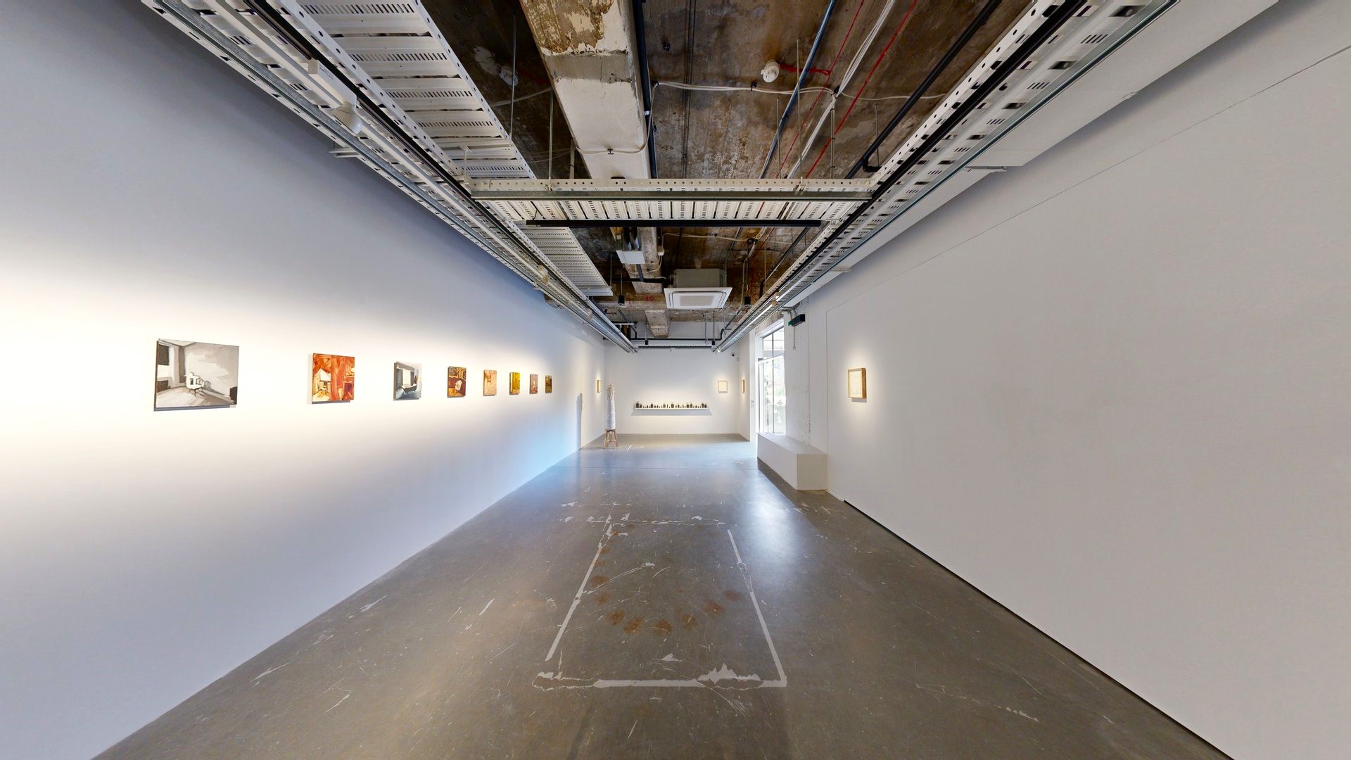 To the left, art paintings decorate the white wall, with another piece on the right. A craft made of ropes and strings is displayed against the far-left wall. The ceiling exposed concrete beams, pipes, air conditioning vents, and pin lights. The polished cement floor displays worn-out markings. To the left, art paintings decorate the white wall, with another piece on the right. A craft made of ropes and strings is displayed against the far-left wall. The ceiling exposed concrete beams, pipes, air conditioning vents, and pin lights. The polished cement floor displays worn-out markings.