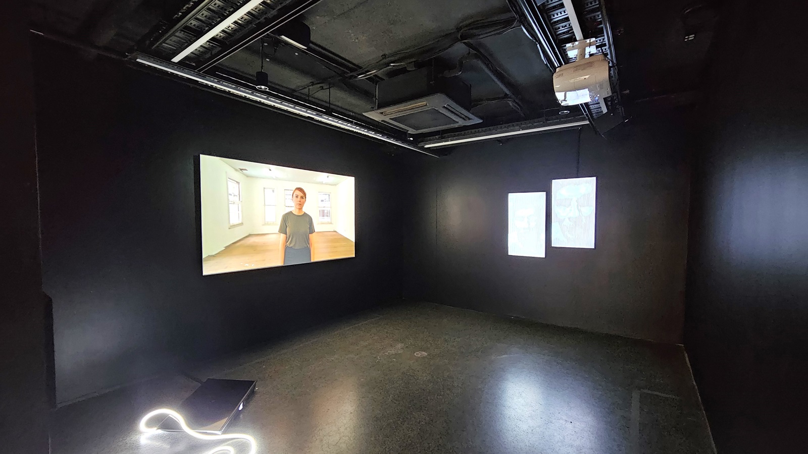 dimly lit room with black tiles, walls, and ceiling. Exposed pipes, a projector, and lighting fixtures adorn the ceiling. A large flatscreen TV is mounted on the left wall. dimly lit room with black tiles, walls, and ceiling. Exposed pipes, a projector, and lighting fixtures adorn the ceiling. A large flatscreen TV is mounted on the left wall.