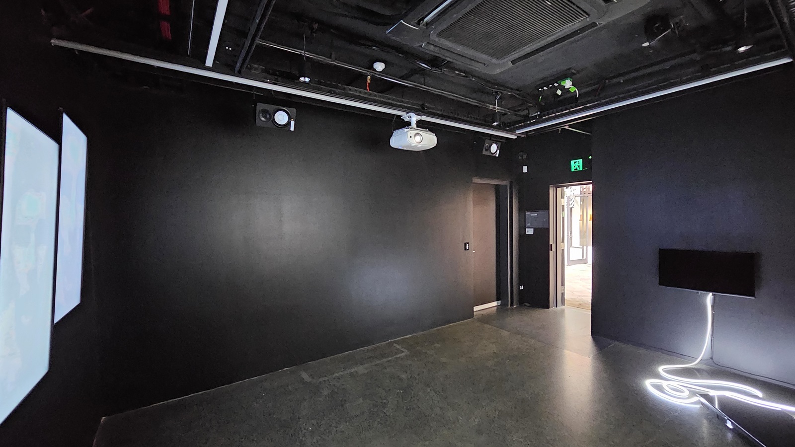 A dimly lit room with black tiles, walls, and ceiling. Exposed pipes, a projector, and lighting fixtures are visible on the ceiling. Digital screens are mounted on the left wall, while a flatscreen is posted to the right. A black door is positioned in the right corner. A dimly lit room with black tiles, walls, and ceiling. Exposed pipes, a projector, and lighting fixtures are visible on the ceiling. Digital screens are mounted on the left wall, while a flatscreen is posted to the right. A black door is positioned in the right corner.