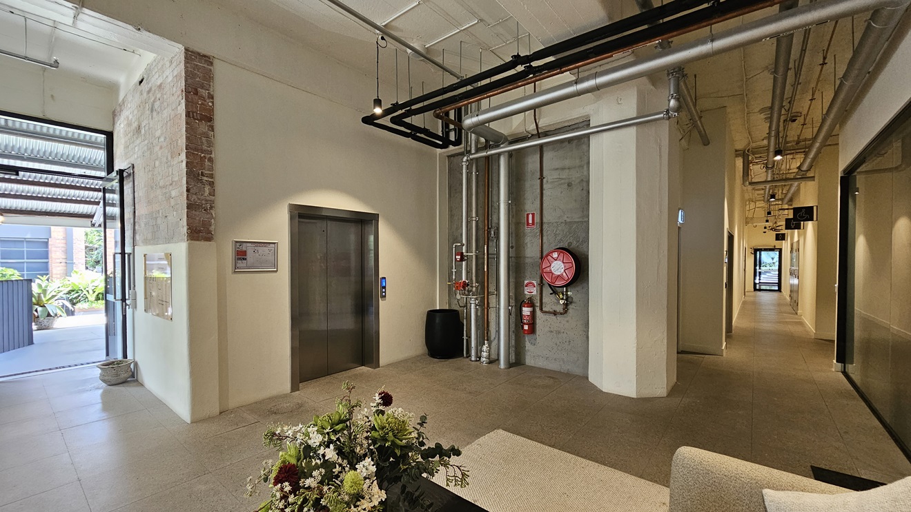 To the left, there's an inward-opening glass door with a small pot nearby. Adjacent is a lift displaying a site map poster. On the concrete wall, a fire extinguisher, fire hose reel, and exposed pipes are mounted. Below is a black rubbish bin. To the right, there's a seating area with a table holding a flower vase. Further in, a corridor leads. The bare ceiling features large pipes, air conditioning vents, and LED lights. To the left, there's an inward-opening glass door with a small pot nearby. Adjacent is a lift displaying a site map poster. On the concrete wall, a fire extinguisher, fire hose reel, and exposed pipes are mounted. Below is a black rubbish bin. To the right, there's a seating area with a table holding a flower vase. Further in, a corridor leads. The bare ceiling features large pipes, air conditioning vents, and LED lights.