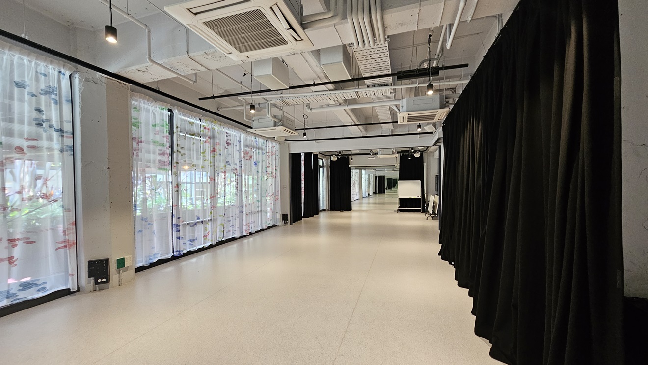 A winding space with white vinyl flooring. To the left, a floor-to-ceiling glass wall with printed white curtains; to the right, black curtains. The ceiling has an industrial look with exposed pipes, air conditioning vents, and LED lights. Foldable tables are visible on the right. A winding space with white vinyl flooring. To the left, a floor-to-ceiling glass wall with printed white curtains; to the right, black curtains. The ceiling has an industrial look with exposed pipes, air conditioning vents, and LED lights. Foldable tables are visible on the right.