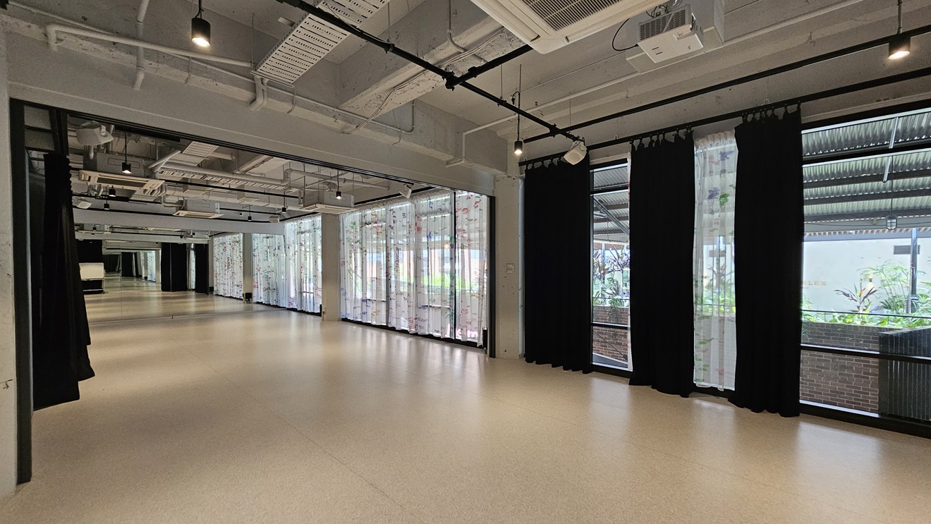 A winding space with white vinyl flooring. To the right, a floor-to-ceiling glass wall with printed white curtains and black curtains. To the left, black curtains hang from the ceiling. The ceiling has an industrial look with exposed pipes, air conditioning vents, and LED lights. A winding space with white vinyl flooring. To the right, a floor-to-ceiling glass wall with printed white curtains and black curtains. To the left, black curtains hang from the ceiling. The ceiling has an industrial look with exposed pipes, air conditioning vents, and LED lights.