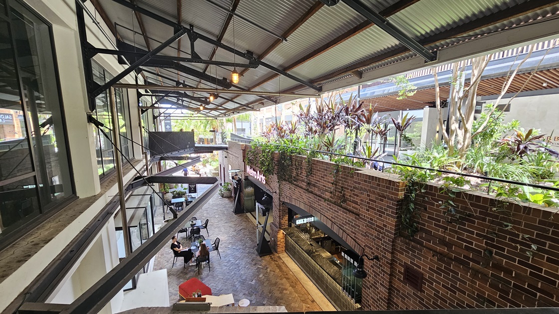 The extended corrugated roofing is supported by large metal beams, with light fixtures hanging from the top. To the right, a landscape garden features ornamental plants hanging from the brick wall. Below, The Metro Arts Bar is visible, with occupied tables and chairs. The extended corrugated roofing is supported by large metal beams, with light fixtures hanging from the top. To the right, a landscape garden features ornamental plants hanging from the brick wall. Below, The Metro Arts Bar is visible, with occupied tables and chairs.