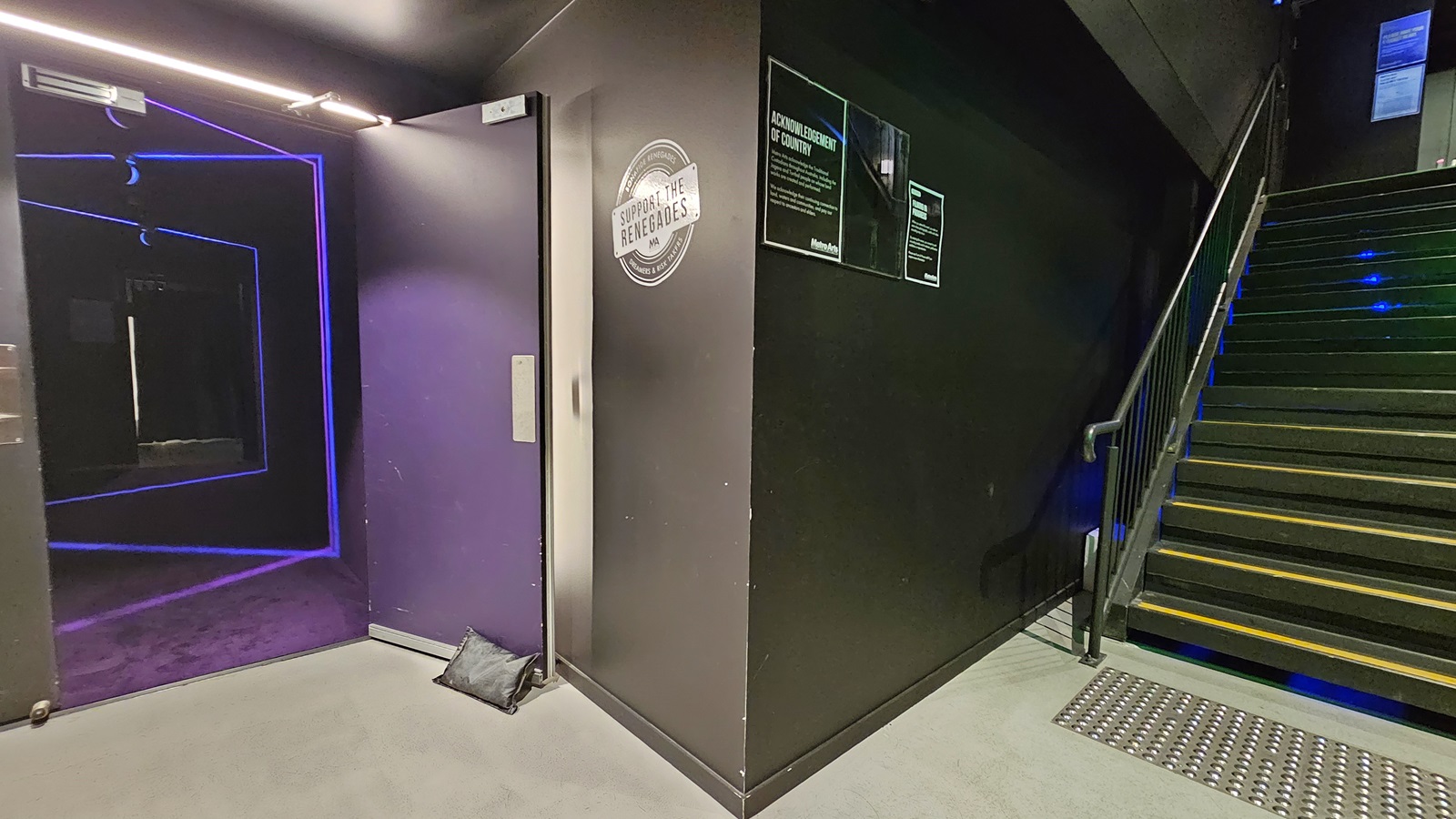 A dimly lit area with black tiles and walls. On the left, a staircase equipped with handrails and truncated domes. The right wall features glowing directional arrows pointing down. The left wall has a rough finish. Further down the stairs, a fire hose reel is visible. A dimly lit area with black tiles and walls. On the left, a staircase equipped with handrails and truncated domes. The right wall features glowing directional arrows pointing down. The left wall has a rough finish. Further down the stairs, a fire hose reel is visible.