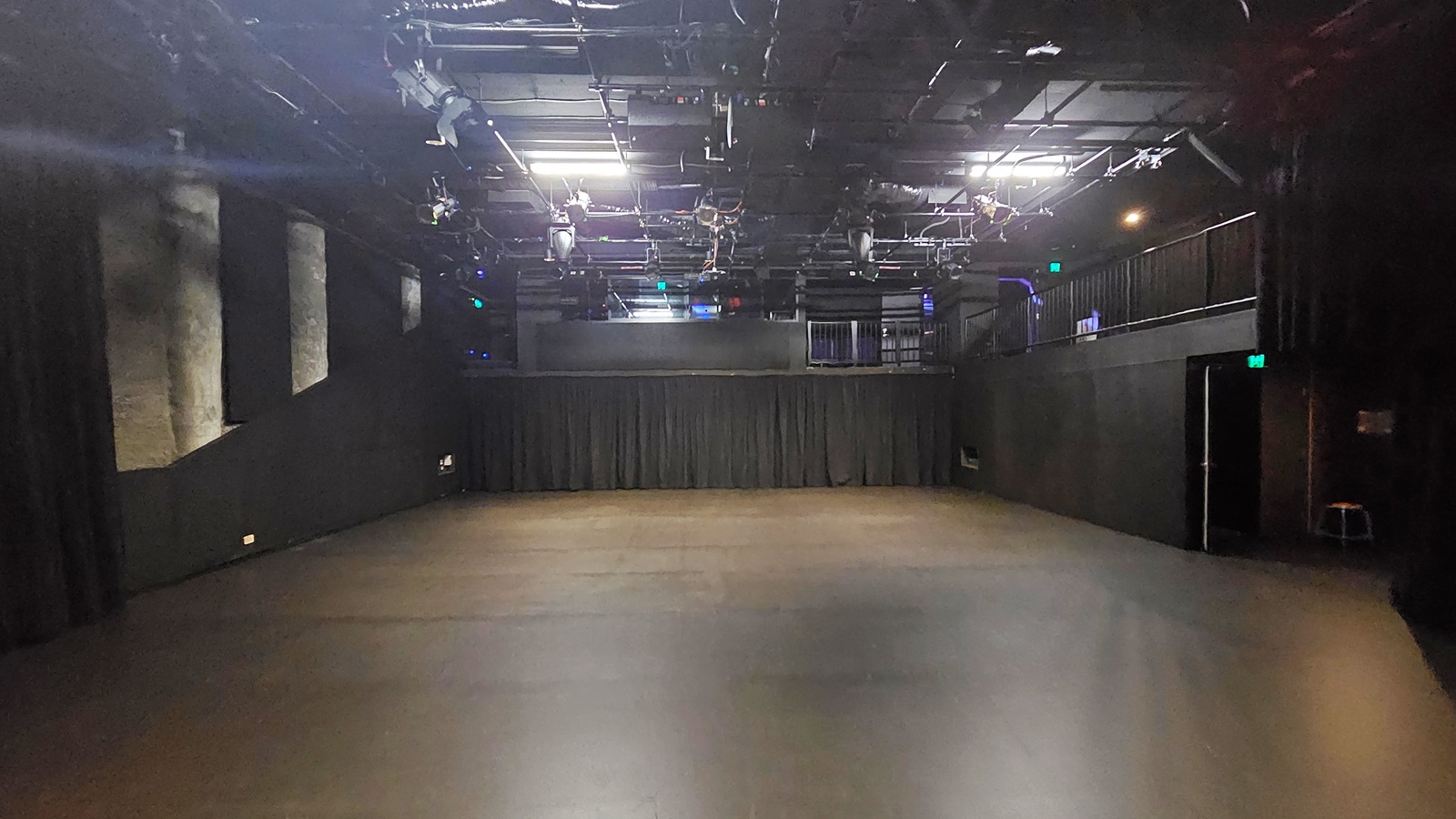 In a dimly lit space with black floor tiles and walls, a door to the right is illuminated by a neon sign reading "Exit". The stage is equipped with black tiles and black curtains. The ceiling is covered with black insulation, hosting audio and light fixtures. To the right, a mezzanine is secured by black metal railings. In a dimly lit space with black floor tiles and walls, a door to the right is illuminated by a neon sign reading "Exit". The stage is equipped with black tiles and black curtains. The ceiling is covered with black insulation, hosting audio and light fixtures. To the right, a mezzanine is secured by black metal railings.