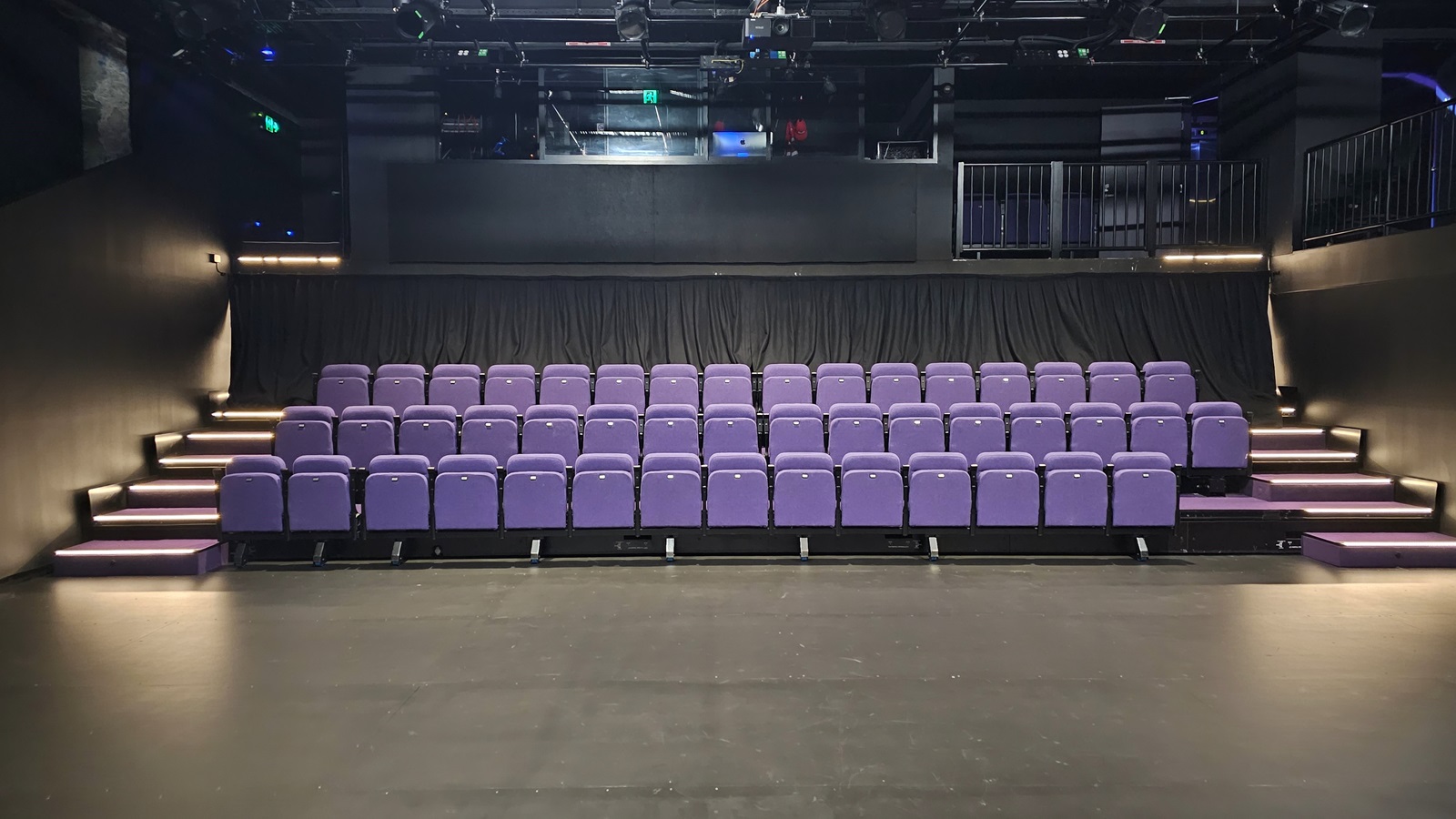 In a dimly lit space with black floor tiles and walls, tiered seating is equipped with lavender chairs. Stairs on both sides lead to the upper tier. The ceiling is covered with black insulation, housing audio, and light fixtures. Behind the seating, a mezzanine secured by black metal railings houses the controls. In a dimly lit space with black floor tiles and walls, tiered seating is equipped with lavender chairs. Stairs on both sides lead to the upper tier. The ceiling is covered with black insulation, housing audio, and light fixtures. Behind the seating, a mezzanine secured by black metal railings houses the controls.