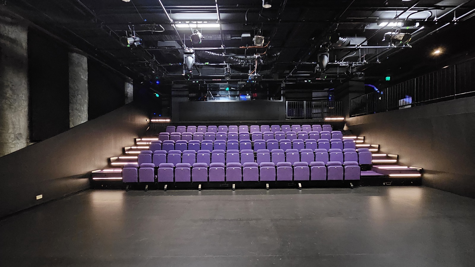 In a dimly lit space with black floor tiles and walls, tiered seating is equipped with lavender chairs. Stairs on both sides lead to the upper tier. The ceiling is covered with black insulation, housing audio, and light fixtures. Behind the seating, a mezzanine secured by black metal railings houses the controls. In a dimly lit space with black floor tiles and walls, tiered seating is equipped with lavender chairs. Stairs on both sides lead to the upper tier. The ceiling is covered with black insulation, housing audio, and light fixtures. Behind the seating, a mezzanine secured by black metal railings houses the controls.