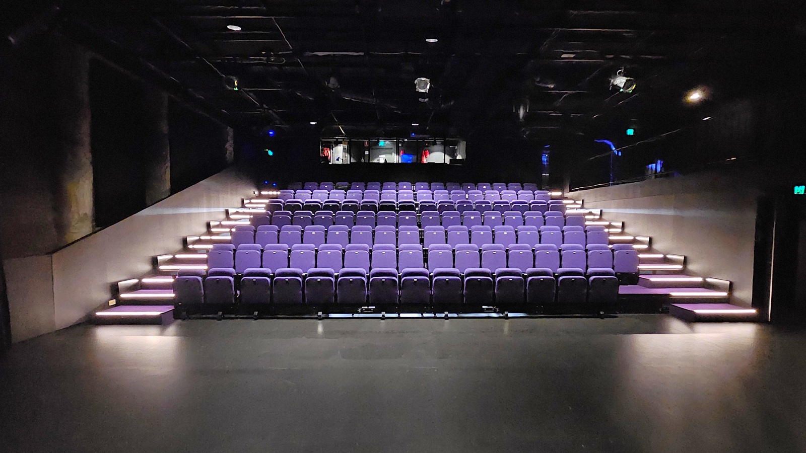 In a dimly lit space with black floor tiles and walls, tiered seating is equipped with lavender chairs. Stairs on both sides lead to the upper tier. The ceiling is covered with black insulation, housing audio, and light fixtures. Behind the seating, a mezzanine secured by black metal railings houses the controls In a dimly lit space with black floor tiles and walls, tiered seating is equipped with lavender chairs. Stairs on both sides lead to the upper tier. The ceiling is covered with black insulation, housing audio, and light fixtures. Behind the seating, a mezzanine secured by black metal railings houses the controls