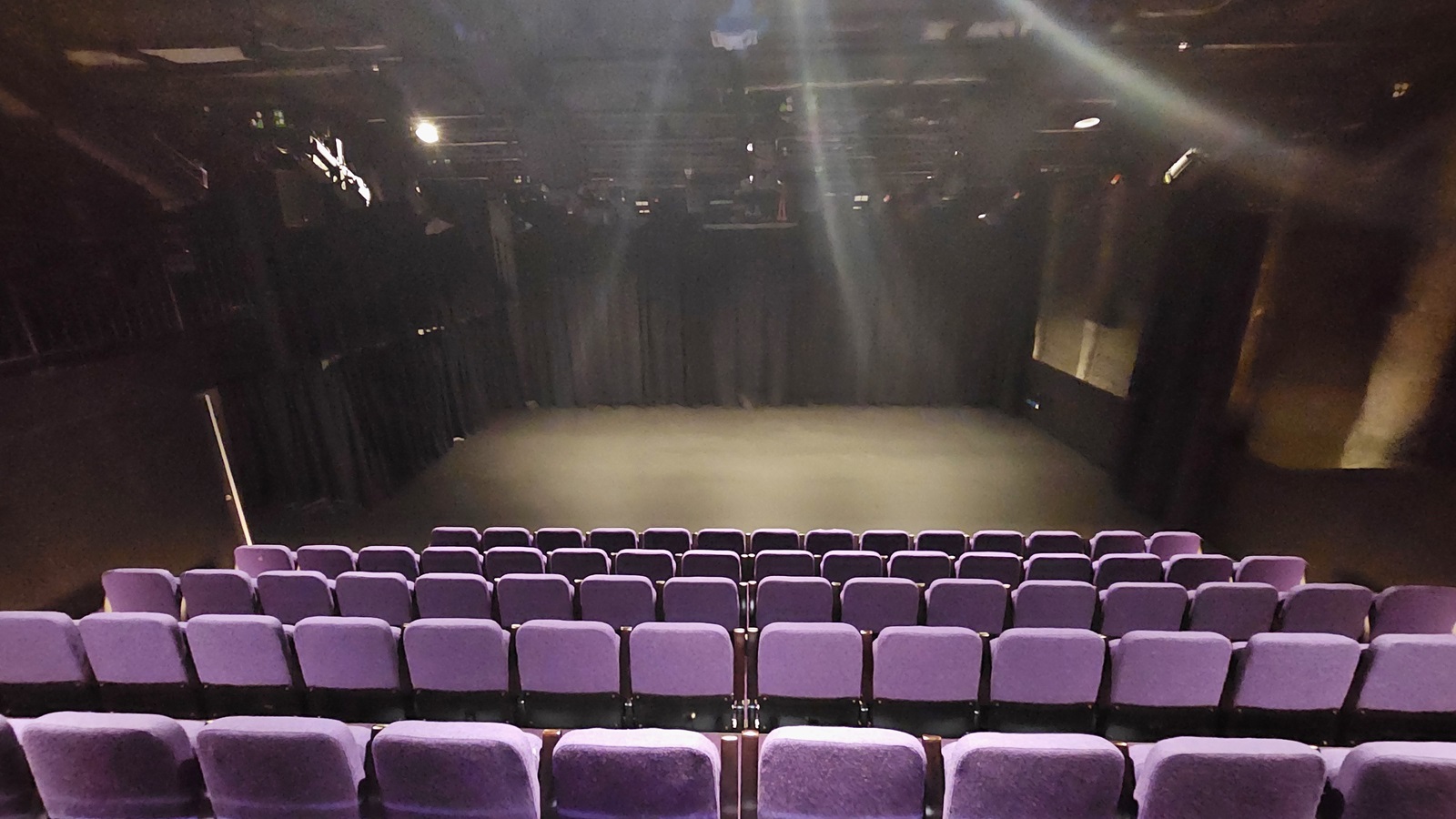 In a dimly lit space with black floor tiles and walls, tiered seating features lavender chairs. The stage in front is adorned with black tiles and surrounded by black walls and curtains. In a dimly lit space with black floor tiles and walls, tiered seating features lavender chairs. The stage in front is adorned with black tiles and surrounded by black walls and curtains.