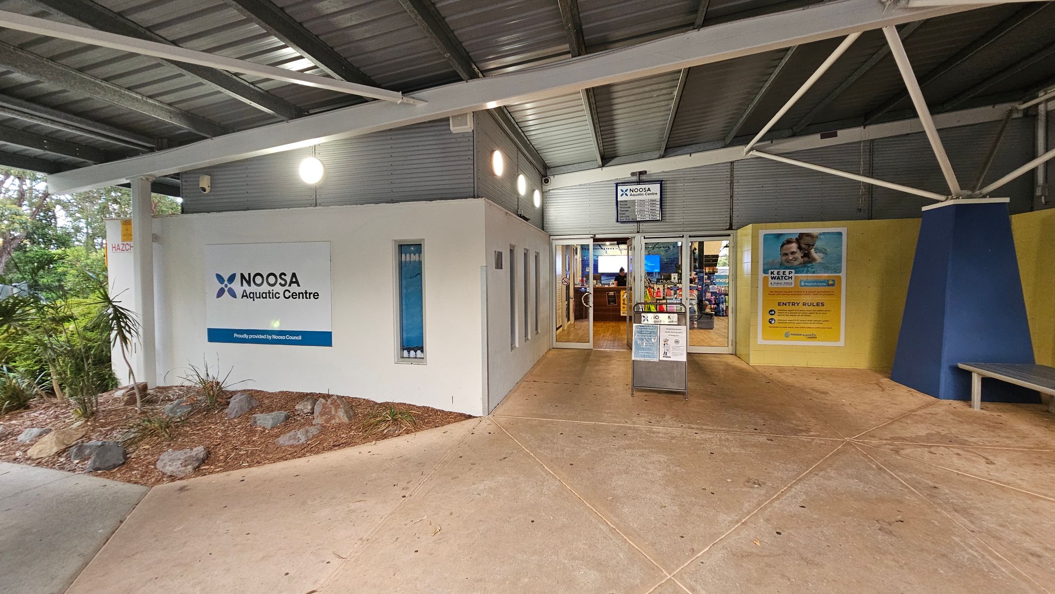 Entrance to the Noosa Aquatic Centre. On the left, a white wall with the centre's sign and a small garden with rocks and plants. In the middle, glass doors lead inside, with a signboard displaying information. On the right, a yellow wall with a poster about entry rules and a blue triangular column. The floor is concrete, and the ceiling is metal with exposed beams.