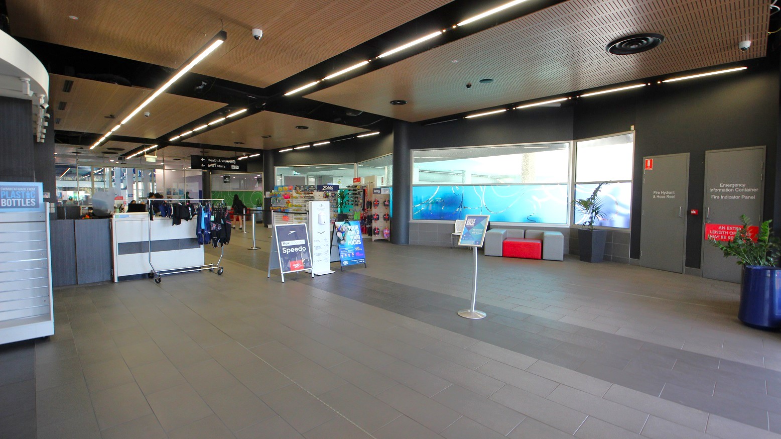A spacious indoor area with a tiled floor and a ceiling featuring linear lights. On the left, there is a display with clothing items and a sign for Speedo. In the centre, there are promotional stands and a seating area with grey and red cushions. The right side has two grey doors with signage, and a large window with a blue aquatic-themed design. A potted plant is visible in the foreground on the right.