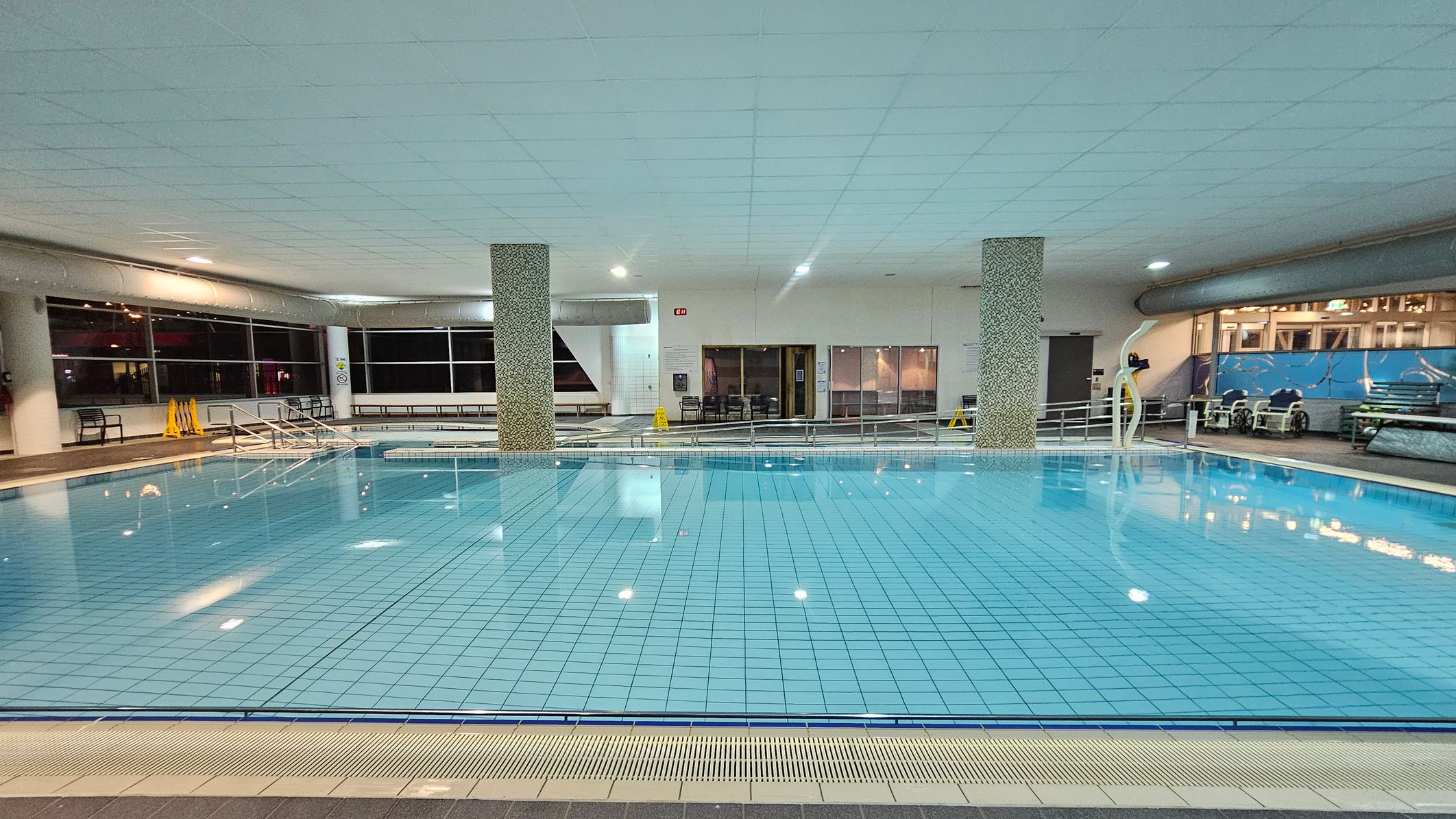 Indoor swimming pool with clear blue water and tiled floor. Two mosaic-covered pillars are in the centre. On the left, large windows and a few benches are visible, with yellow caution signs nearby. The right side has a mural and some poolside equipment. The ceiling is white with recessed lighting, and the pool area is surrounded by a tiled walkway.