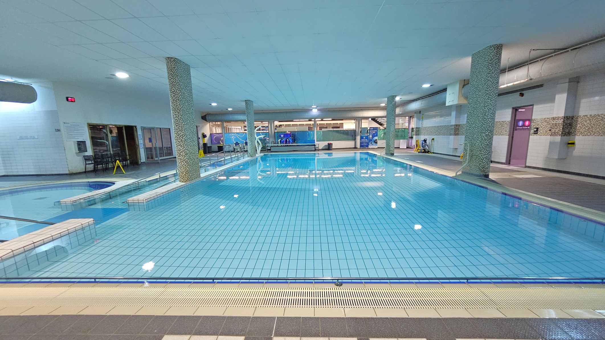 An indoor swimming pool with clear blue water and tiled flooring. Several mosaic-tiled columns are positioned around the pool. On the left, there is a smaller, circular pool area. The background features large windows and doors, with a staircase visible on the right. The ceiling is white with recessed lighting, and the floor surrounding the pool is tiled in shades of grey and beige. A caution sign is placed near the entrance on the left.