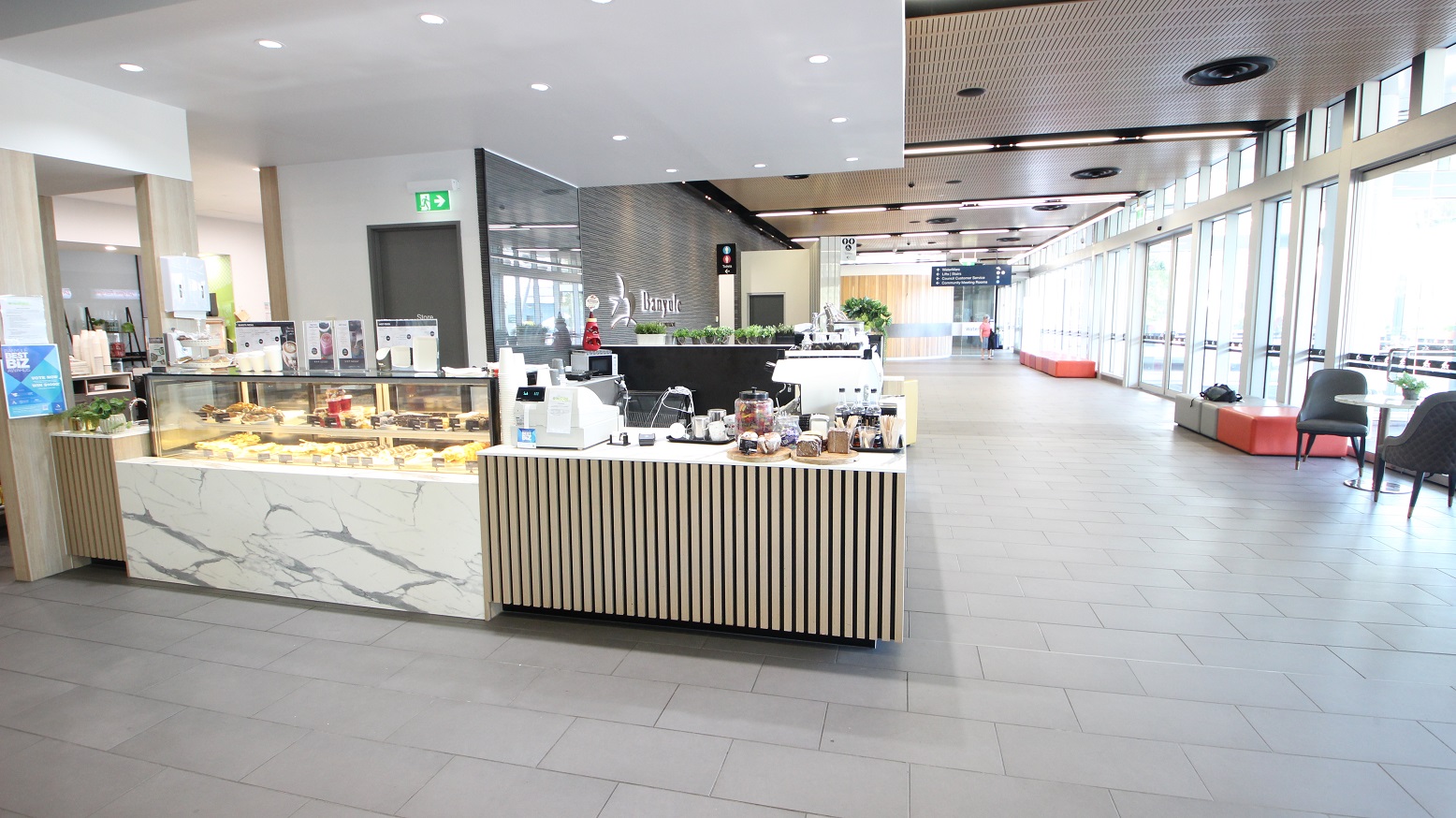 A modern café interior with a marble and wood counter on the left displaying pastries and a coffee machine. The floor is tiled in grey, and the ceiling has recessed lighting. On the right, large windows allow natural light to flood in, with seating areas featuring grey chairs and orange benches. In the background, a corridor leads to other areas, with signs and more seating visible.