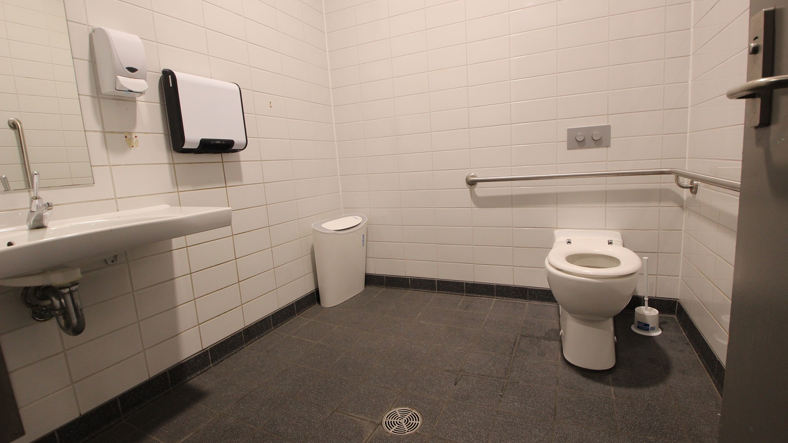 A public restroom with white tiled walls and a grey tiled floor. On the left, a wall-mounted sink with a soap dispenser and a paper towel holder above it. In the middle, a white waste bin is placed against the wall. On the right, a toilet with a grab bar beside it and a toilet brush on the floor. A metal door handle is visible on the right edge.