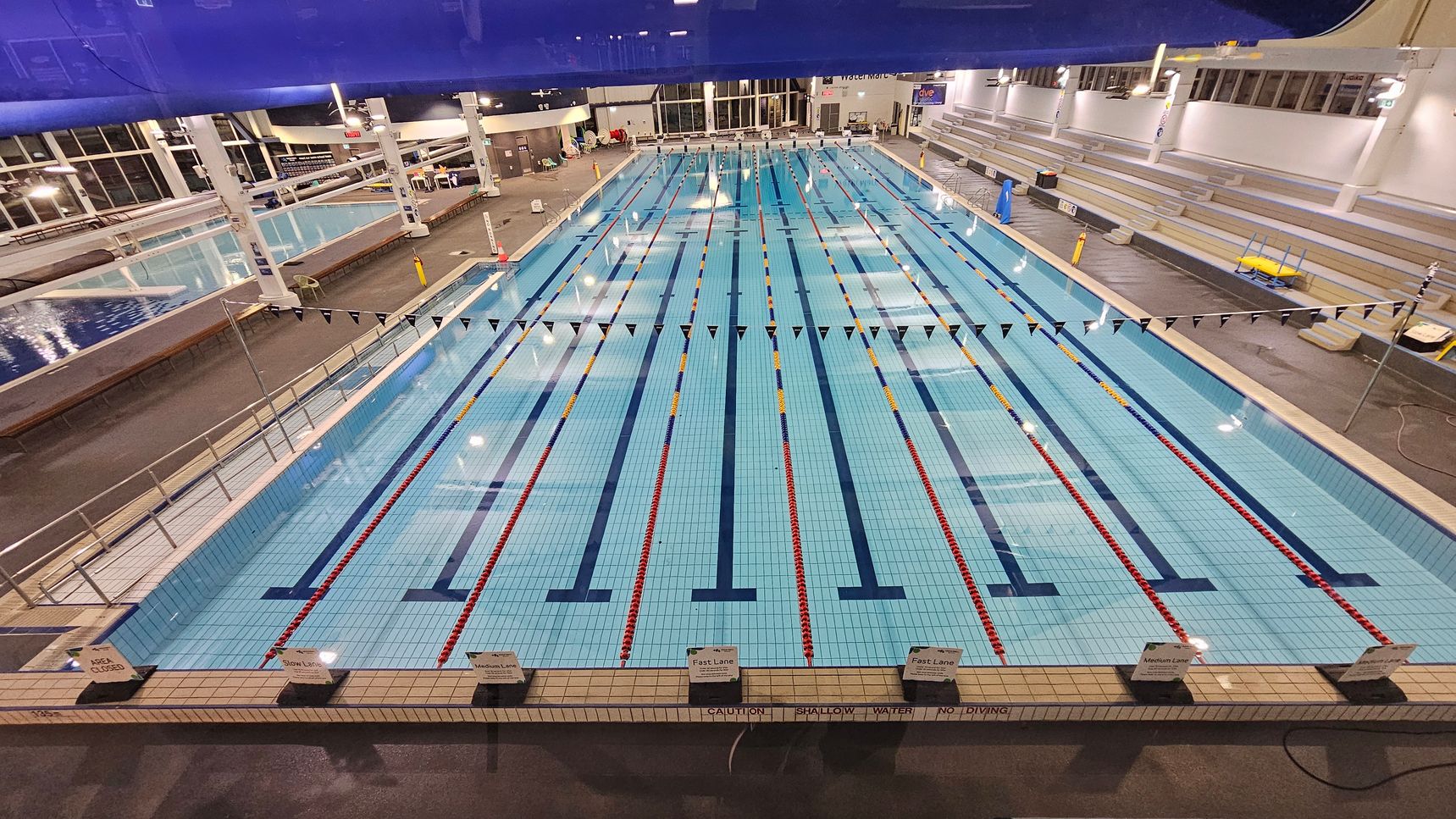 An indoor swimming pool with multiple lanes, each marked by red and yellow lane dividers. Overhead, a row of triangular flags stretches across the pool. On the right, there are tiered seating areas with benches. To the left, another smaller pool is visible. The floor surrounding the pool is dark and wet, and the ceiling is high with exposed beams and lighting fixtures. Signs at the pool's edge indicate lane speeds.