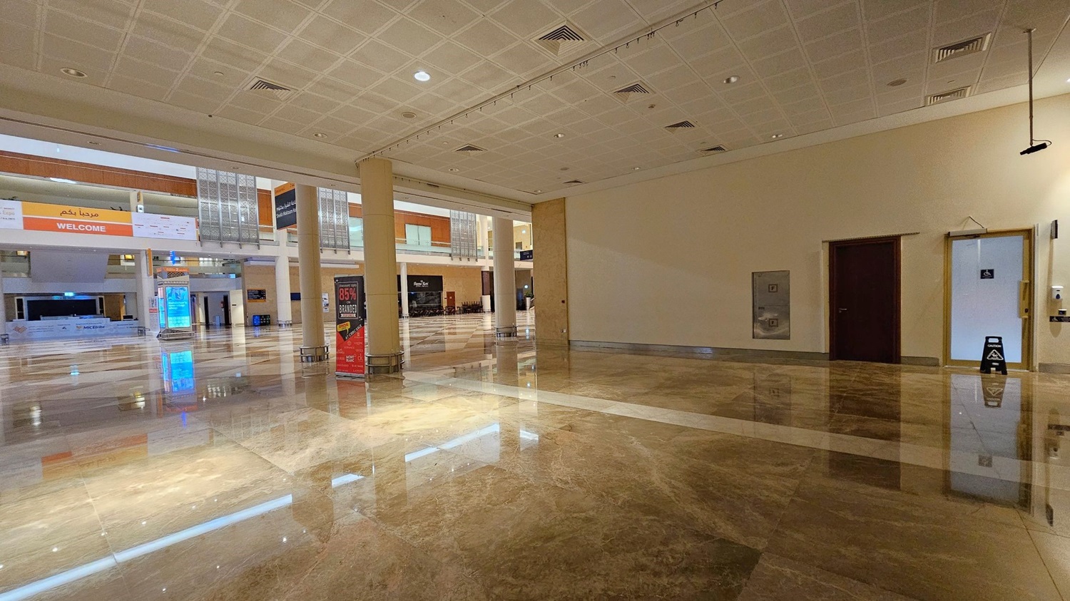 A spacious indoor area with a shiny marble floor. On the left, there's a door with a disability sign and a wall-mounted hand sanitiser dispenser. In the centre, a small white partitioned area is set up. To the right, a large open space with columns and a colourful sign advertising a sale. In the background, a second level with wooden panels and a "WELCOME" banner is visible. The ceiling is tiled with recessed lighting. A spacious indoor area with a shiny marble floor. On the left, there's a door with a disability sign and a wall-mounted hand sanitiser dispenser. In the centre, a small white partitioned area is set up. To the right, a large open space with columns and a colourful sign advertising a sale. In the background, a second level with wooden panels and a "WELCOME" banner is visible. The ceiling is tiled with recessed lighting.