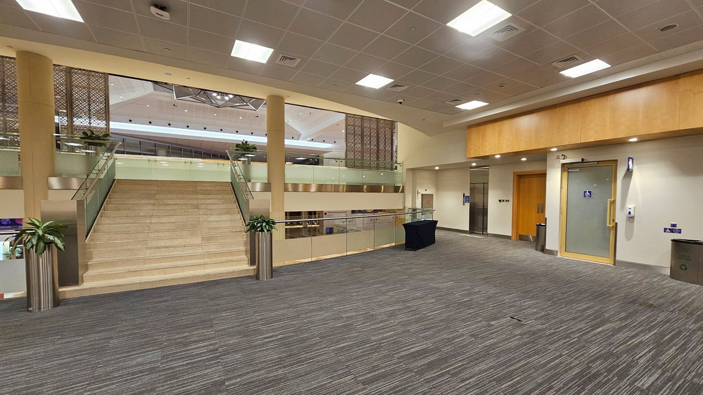 A spacious indoor area with a wide staircase in the centre, flanked by glass railings and potted plants. The floor is covered with grey and green striped carpet. On the right, there is a wooden door and a glass door with a disabled access sign. The ceiling is fitted with square lights and a grid pattern. In the background, there are decorative panels and a glimpse of an upper level with more plants. A spacious indoor area with a wide staircase in the centre, flanked by glass railings and potted plants. The floor is covered with grey and green striped carpet. On the right, there is a wooden door and a glass door with a disabled access sign. The ceiling is fitted with square lights and a grid pattern. In the background, there are decorative panels and a glimpse of an upper level with more plants.
