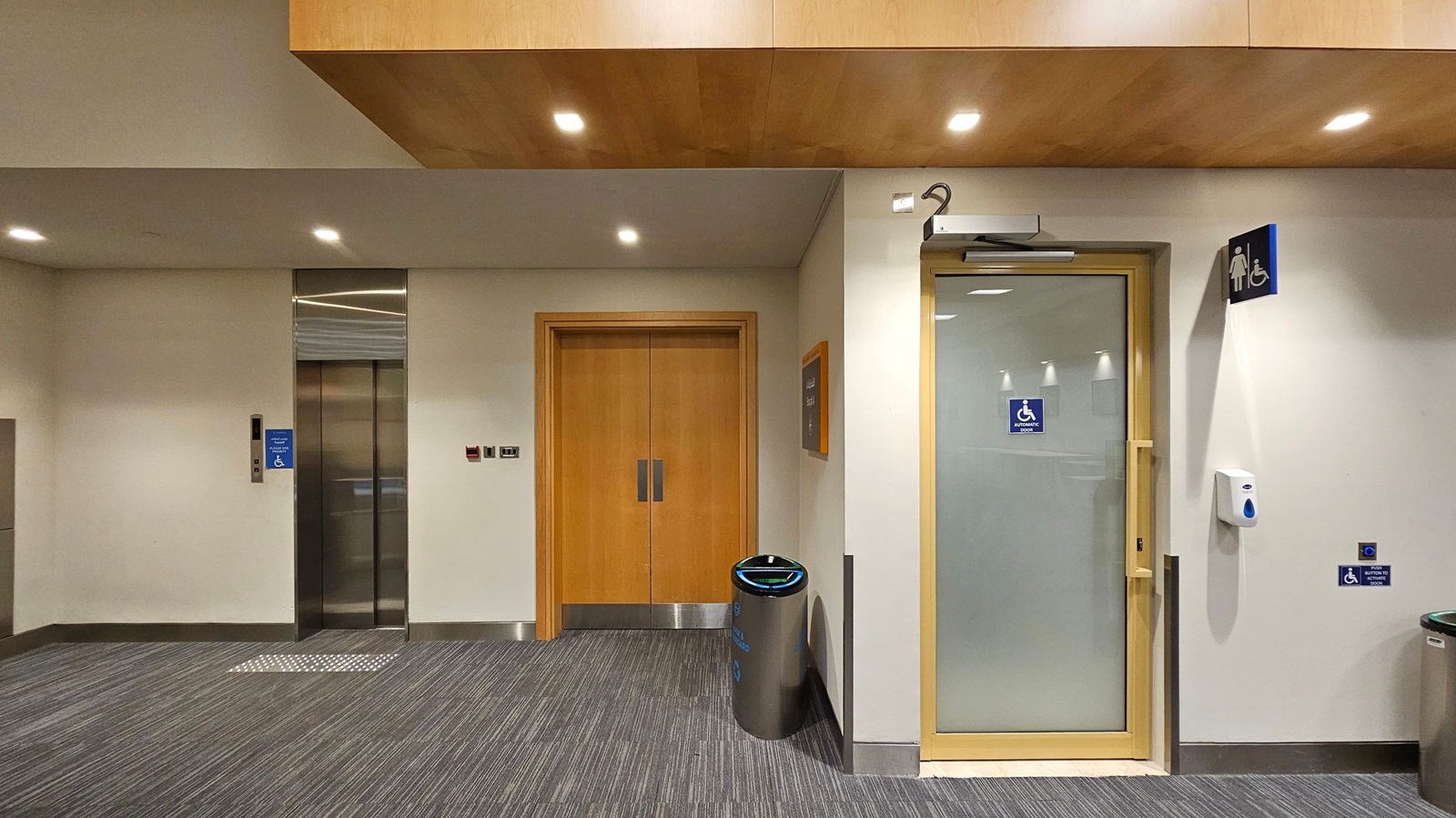 A hallway with a grey carpeted floor and a light-coloured ceiling with recessed lighting. On the left, there is a lift with a blue accessibility sign. In the centre, a wooden double door is flanked by a metal bin. On the right, a door with a glass panel has a sign for accessible toilets. Above it, a women's toilet sign is visible. A hand sanitiser dispenser is mounted on the wall next to the door. A hallway with a grey carpeted floor and a light-coloured ceiling with recessed lighting. On the left, there is a lift with a blue accessibility sign. In the centre, a wooden double door is flanked by a metal bin. On the right, a door with a glass panel has a sign for accessible toilets. Above it, a women's toilet sign is visible. A hand sanitiser dispenser is mounted on the wall next to the door.