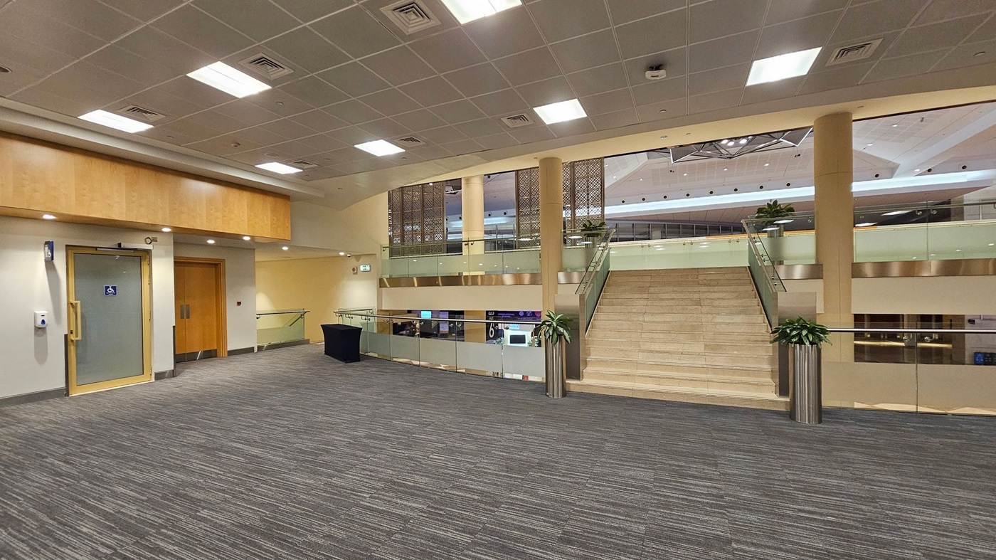 A spacious indoor area with a grey carpeted floor. On the left, there is a door with a disabled access sign and a wooden double door. In the centre, a wide staircase with glass railings leads to an upper level. Potted plants are placed at the base and top of the stairs. The ceiling is fitted with square lights and ventilation panels. The upper level has a decorative panel and overlooks a lower area with visible signage. A spacious indoor area with a grey carpeted floor. On the left, there is a door with a disabled access sign and a wooden double door. In the centre, a wide staircase with glass railings leads to an upper level. Potted plants are placed at the base and top of the stairs. The ceiling is fitted with square lights and ventilation panels. The upper level has a decorative panel and overlooks a lower area with visible signage.