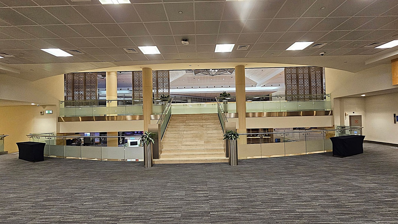A spacious indoor area with a wide staircase in the centre leading to an upper level. The staircase is flanked by glass railings with metal accents. On either side of the staircase are large potted plants. The floor is carpeted in a grey and green pattern. The ceiling is fitted with square panels and recessed lighting. On the left and right, there are black covered tables. The background features decorative panels and a glimpse of a ceiling with geometric patterns. A spacious indoor area with a wide staircase in the centre leading to an upper level. The staircase is flanked by glass railings with metal accents. On either side of the staircase are large potted plants. The floor is carpeted in a grey and green pattern. The ceiling is fitted with square panels and recessed lighting. On the left and right, there are black covered tables. The background features decorative panels and a glimpse of a ceiling with geometric patterns.