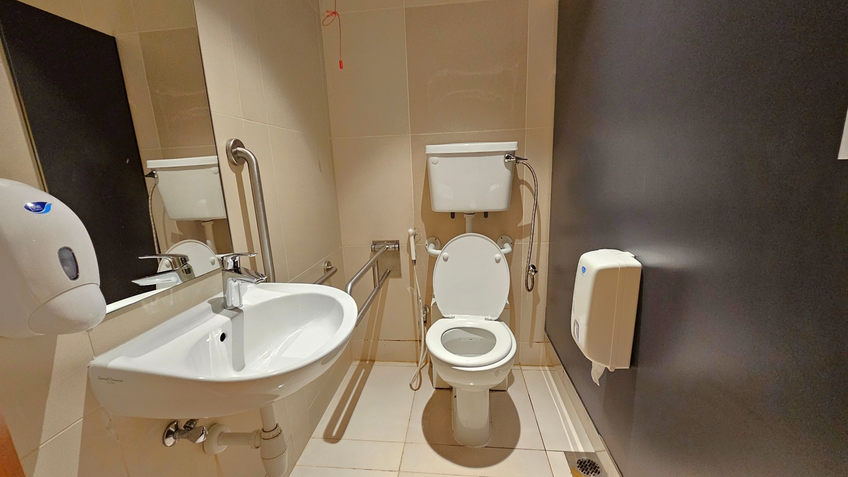 A bathroom with beige tiled walls and floor. On the left, a white sink with a chrome tap is mounted on the wall, accompanied by a soap dispenser and a large mirror. In the centre, a toilet with a white cistern and a handheld bidet spray is installed. Stainless steel grab bars are positioned around the toilet. On the right, a toilet paper dispenser is attached to a dark grey wall. A red emergency pull cord hangs from the ceiling. A bathroom with beige tiled walls and floor. On the left, a white sink with a chrome tap is mounted on the wall, accompanied by a soap dispenser and a large mirror. In the centre, a toilet with a white cistern and a handheld bidet spray is installed. Stainless steel grab bars are positioned around the toilet. On the right, a toilet paper dispenser is attached to a dark grey wall. A red emergency pull cord hangs from the ceiling.
