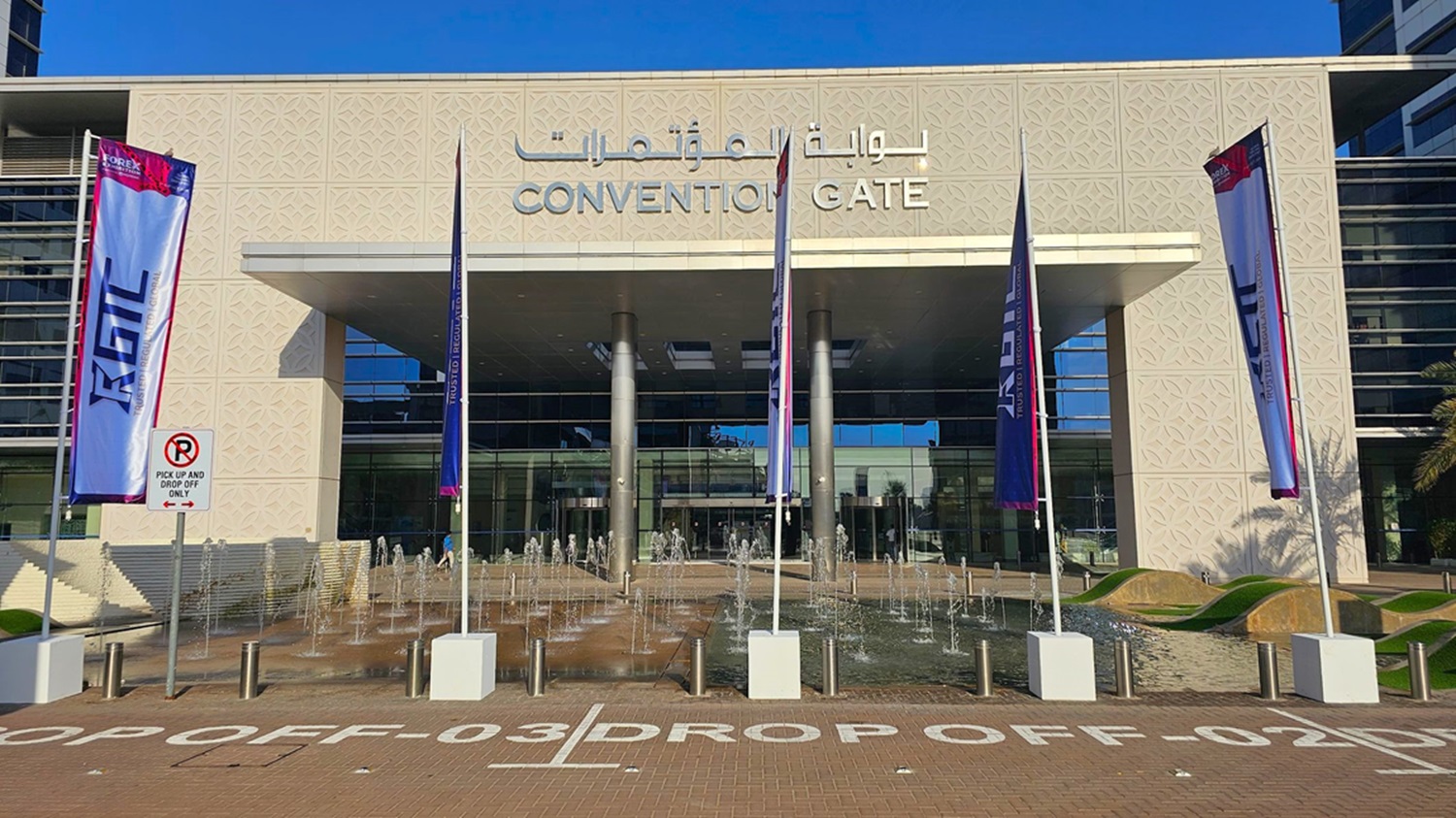 A modern building with a glass facade on the right, featuring large windows and a sleek design. On the left, there are palm trees lining a paved walkway. The building has a decorative patterned wall and a large overhang supported by metal columns. The ground is covered with interlocking bricks, and there are bollards along the path. Shadows of the palm trees are cast on the ground. A modern building with a glass facade on the right, featuring large windows and a sleek design. On the left, there are palm trees lining a paved walkway. The building has a decorative patterned wall and a large overhang supported by metal columns. The ground is covered with interlocking bricks, and there are bollards along the path. Shadows of the palm trees are cast on the ground.