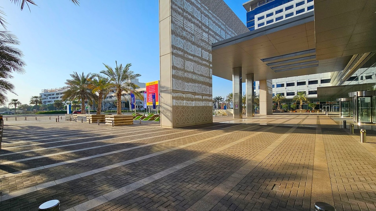 A modern outdoor plaza with a patterned, reflective building on the right. The ground is paved with alternating light and dark bricks. Palm trees and benches are on the left, with a large, colourful sign in the background. The area is open and spacious, with a clear blue sky above. A glass entrance with revolving doors is visible on the right side of the building. A modern outdoor plaza with a patterned, reflective building on the right. The ground is paved with alternating light and dark bricks. Palm trees and benches are on the left, with a large, colourful sign in the background. The area is open and spacious, with a clear blue sky above. A glass entrance with revolving doors is visible on the right side of the building.