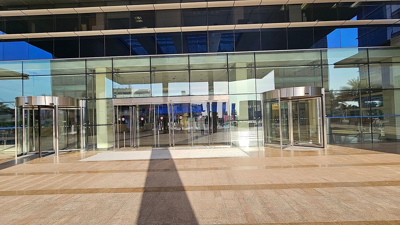A modern building entrance with large glass doors and windows. Two revolving doors are on the left and right, framed in metallic silver. The central section features multiple glass sliding doors. The floor is tiled in a pattern of light and dark brown. Reflections of the surrounding area, including trees and other structures, are visible in the glass. The ceiling inside is white with recessed lighting. A modern building entrance with large glass doors and windows. Two revolving doors are on the left and right, framed in metallic silver. The central section features multiple glass sliding doors. The floor is tiled in a pattern of light and dark brown. Reflections of the surrounding area, including trees and other structures, are visible in the glass. The ceiling inside is white with recessed lighting.